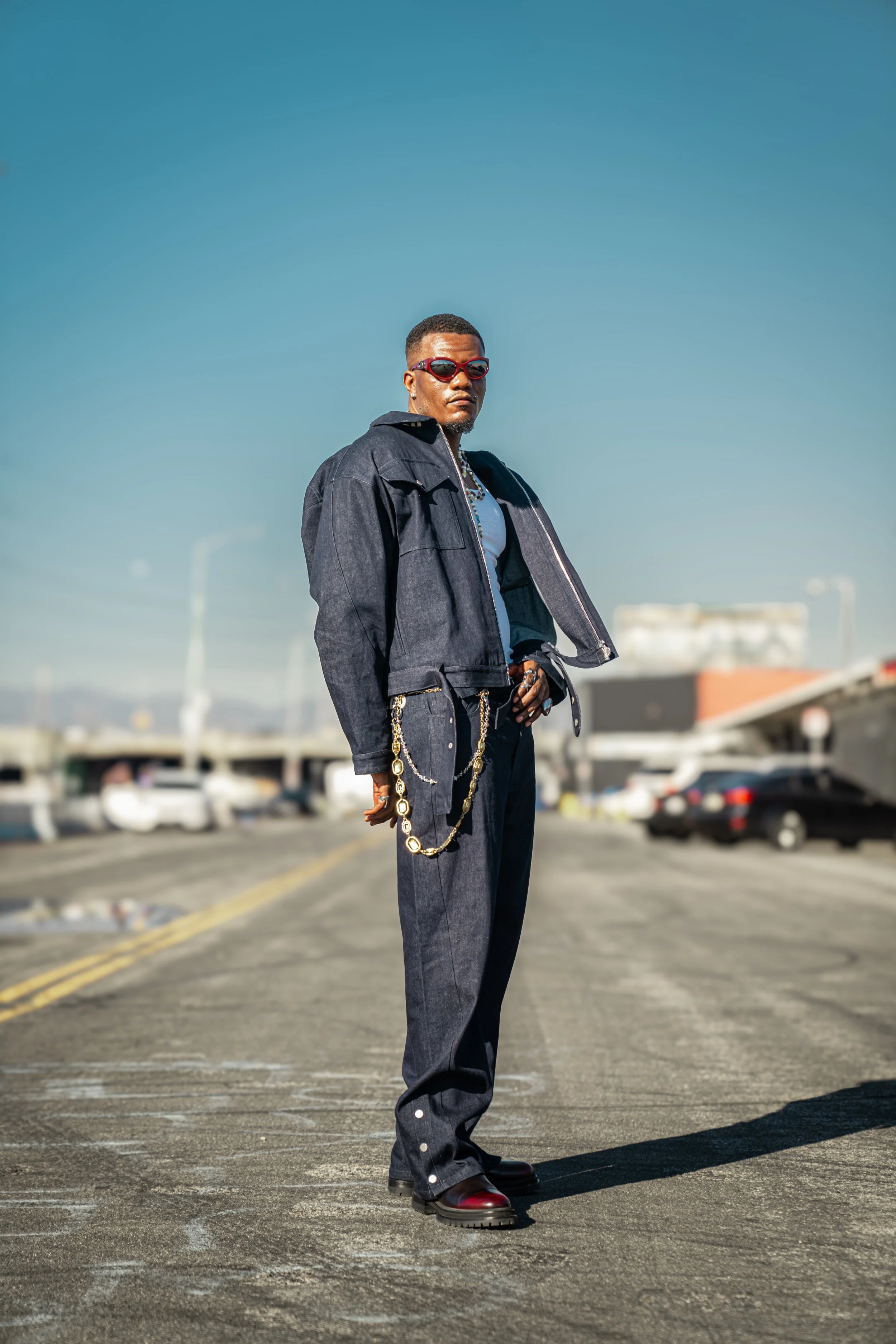 Man standing on an empty parking lot, wearing a dark denim jacket and pants, with red sunglasses, jewelry, and boots, with a clear blue sky in the background.