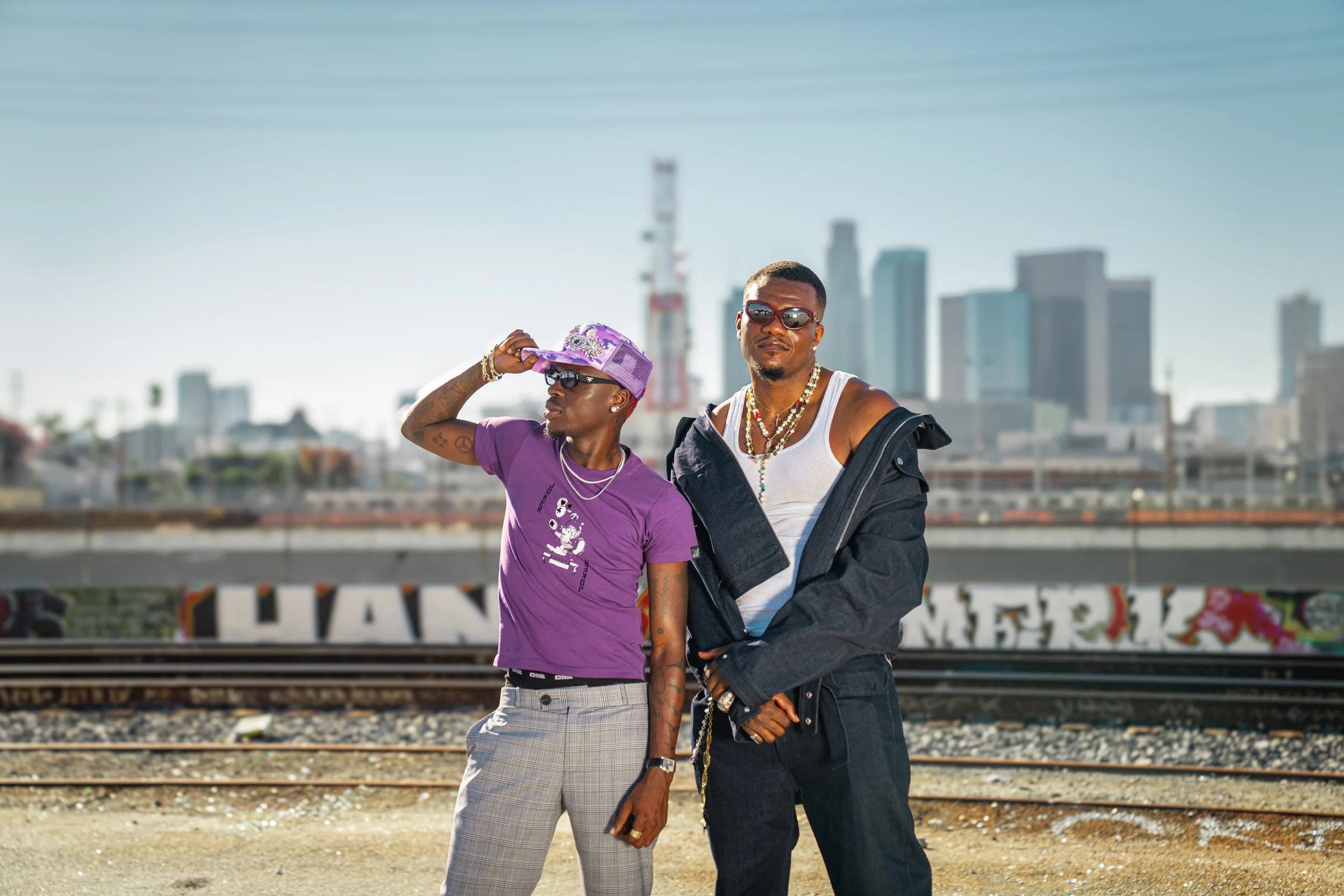 Two stylish men standing on a railroad track in an urban area with a city skyline in the background. One man is wearing a purple t-shirt, plaid pants, a purple hat, and sunglasses, while the other is wearing a white tank top, a black jacket off the shoulders, and sunglasses. They are accessorized with jewelry.