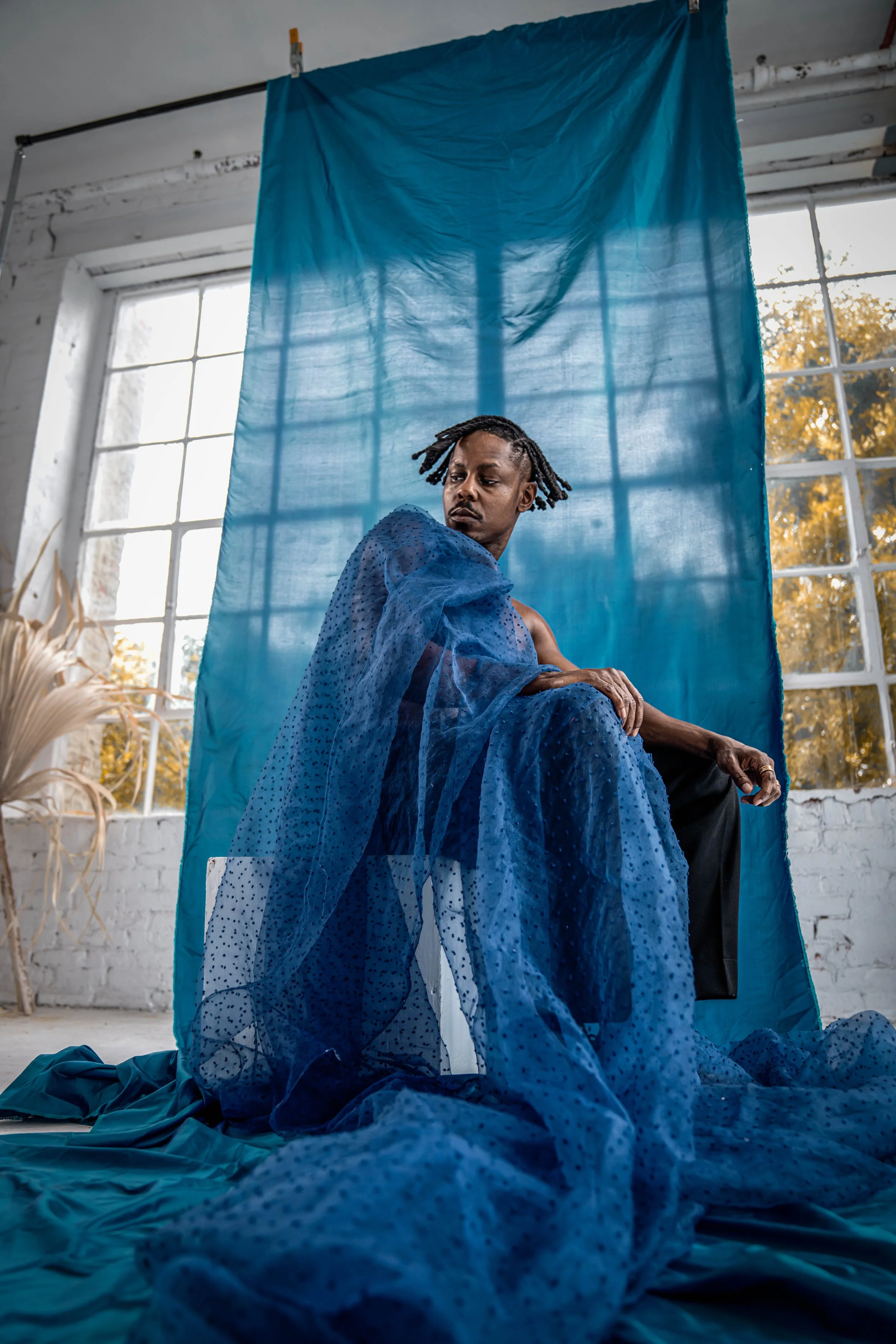 A person sitting in a room with large windows, draped in a sheer blue fabric with a dotted pattern, with a blue backdrop behind them and sunlight coming through the windows.