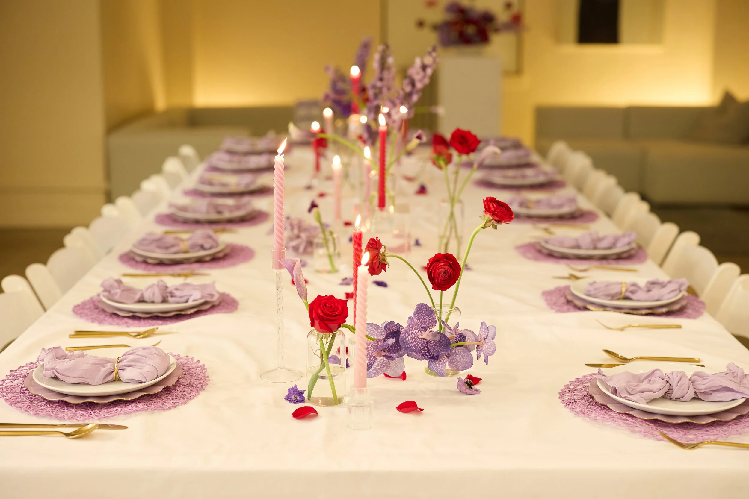 A long dining table set for a celebration with purple, pink, and red flowers, pink candles, and lavender napkins. The table has white plates, gold utensils, and doilies under the plates, arranged in a symmetrical pattern.