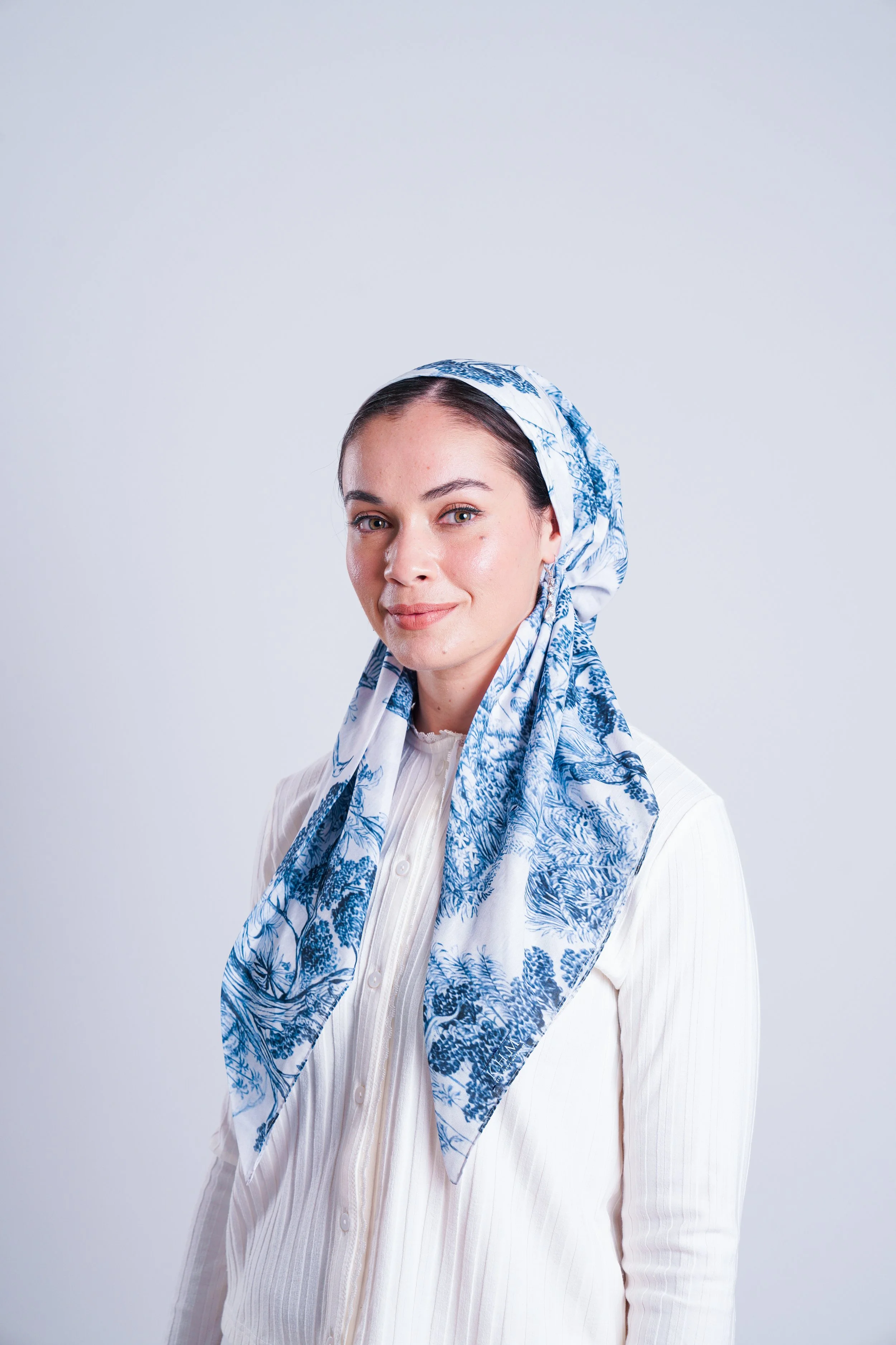 A woman with light skin and dark hair pulled back, wearing a white blouse and a blue and white patterned headscarf, stands against a plain light background and looks at the camera with a slight smile.