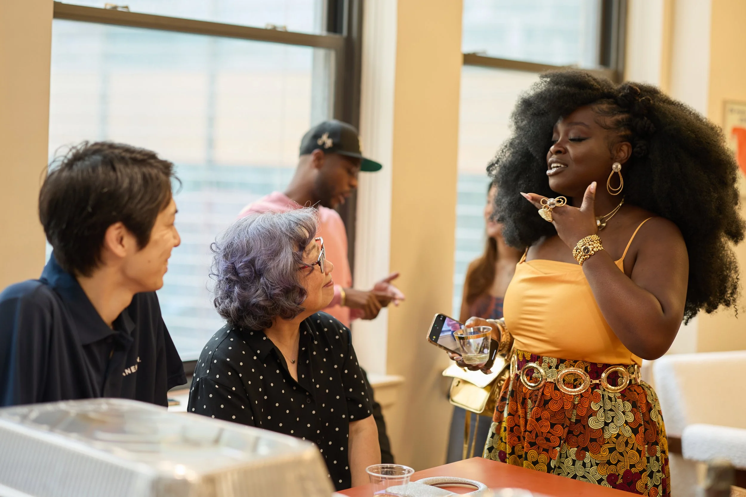 A woman in a yellow top and patterned skirt, wearing gold jewelry, talking to three seated women at a social gathering indoors near windows.
