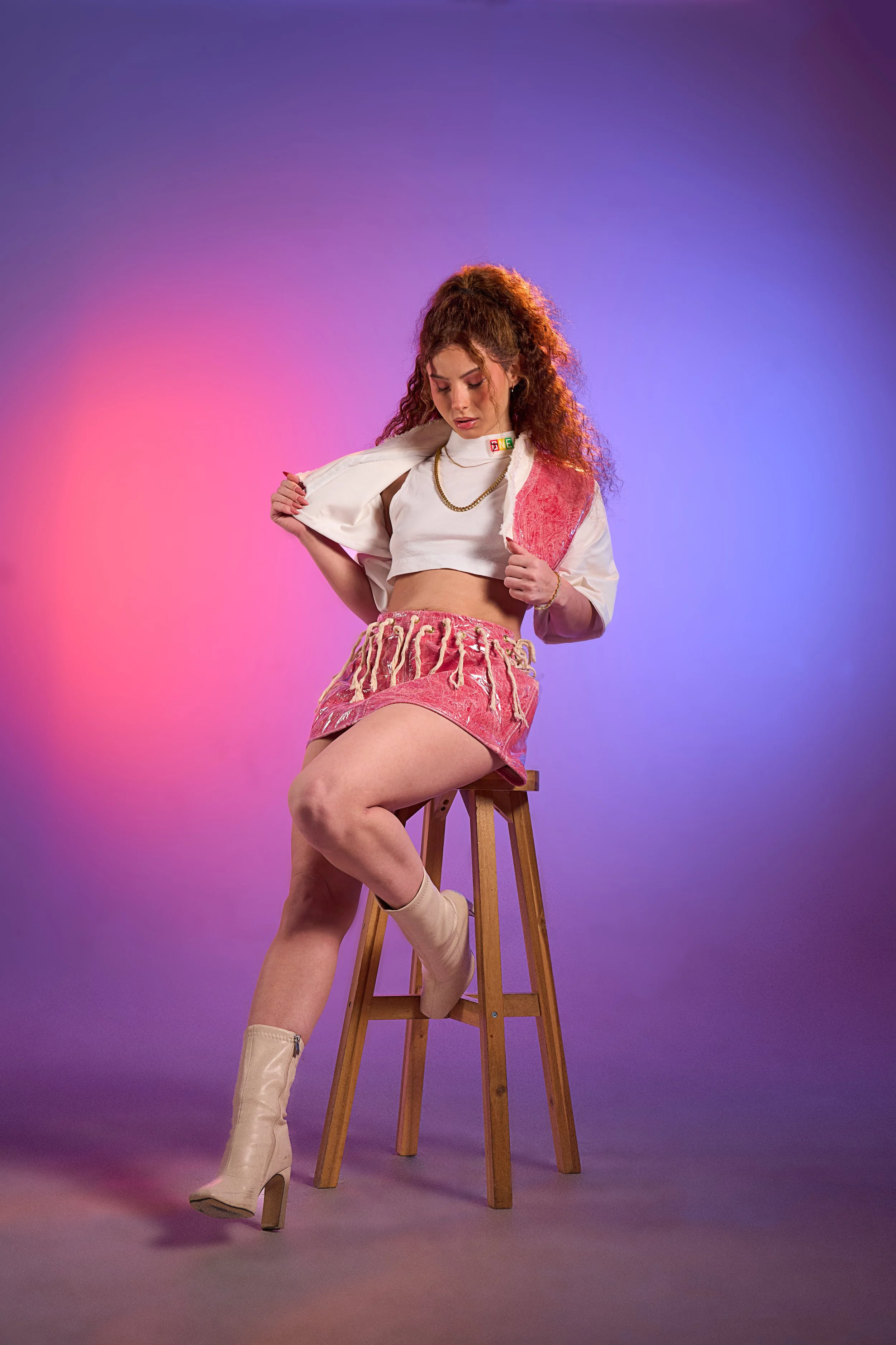 A young woman with curly red hair sits on a wooden stool against a gradient background of pink and purple hues. She is wearing a white cropped top, a pink velvet jacket, a pink skirt with a textured design, beige heeled boots, and minimal jewelry.