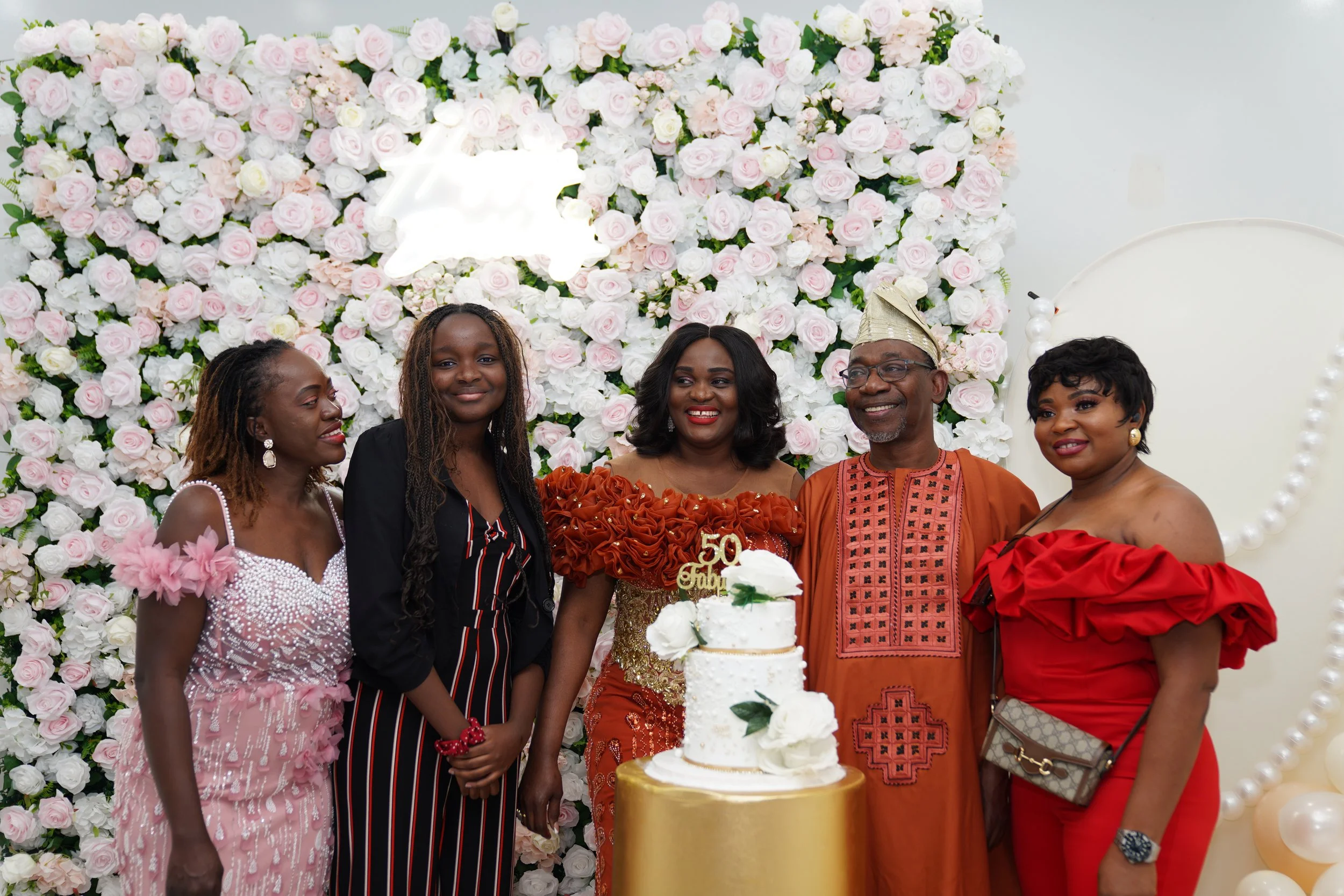 A group of six people celebrating a 50th birthday in front of a floral backdrop with pink and white roses. There is a tiered white cake with white flowers and a gold cake topper reading '50 Gbami'.