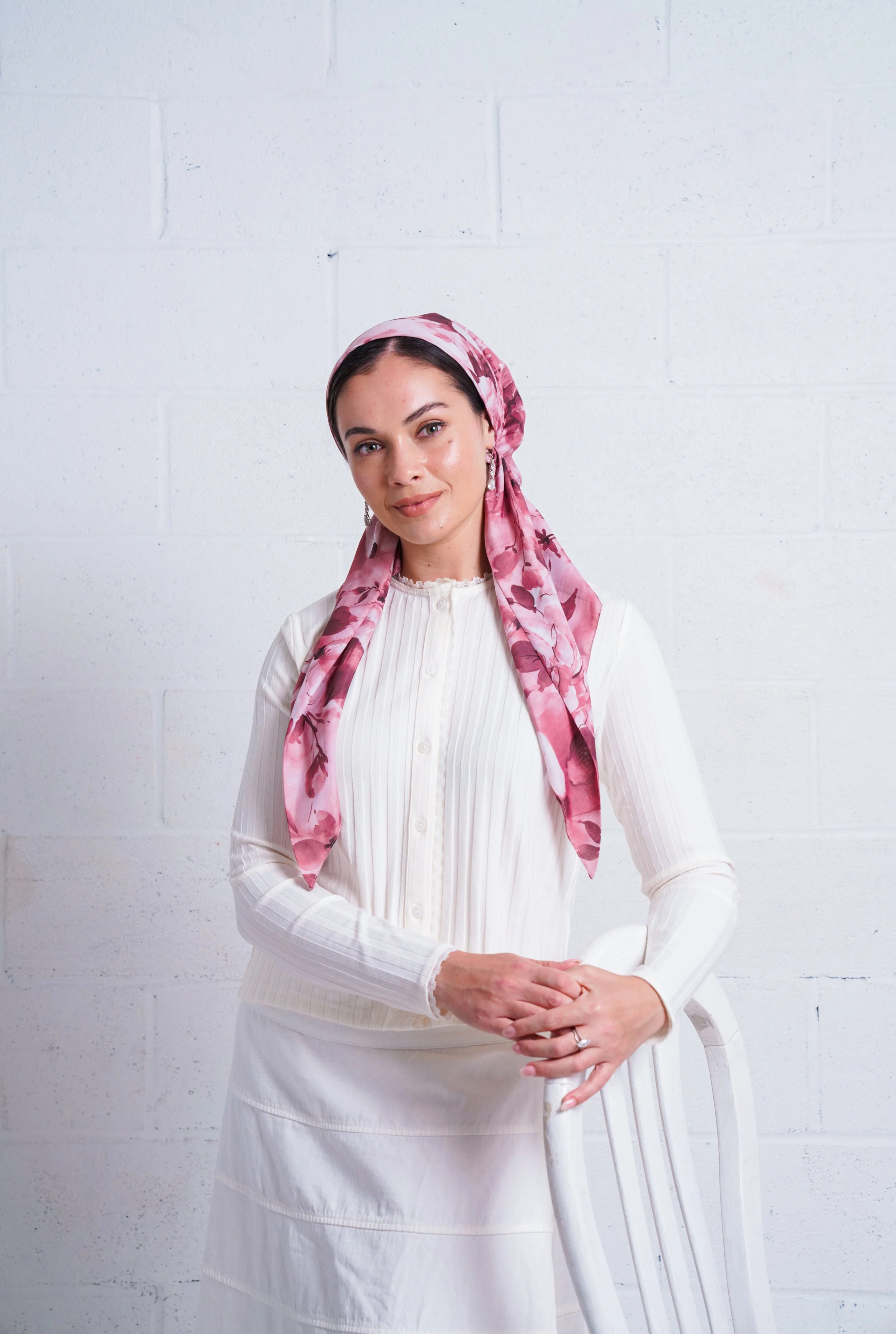 A woman with a pink floral headscarf, wearing a white button-up top and skirt, leaning on a chair against a white brick wall.