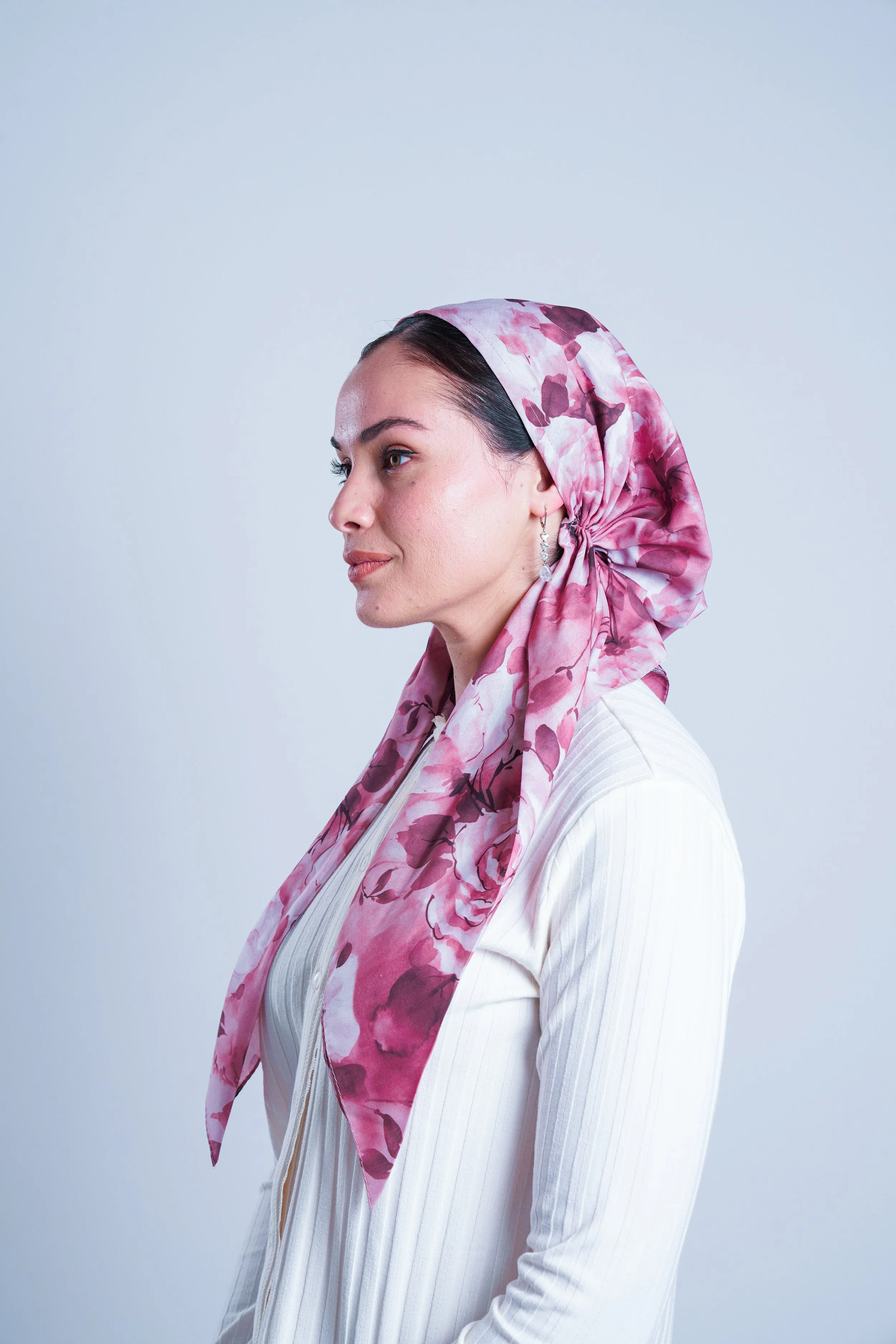 A woman with dark hair, wearing a pink floral headscarf, a white ribbed top, and pearl earrings, is in profile against a plain background.