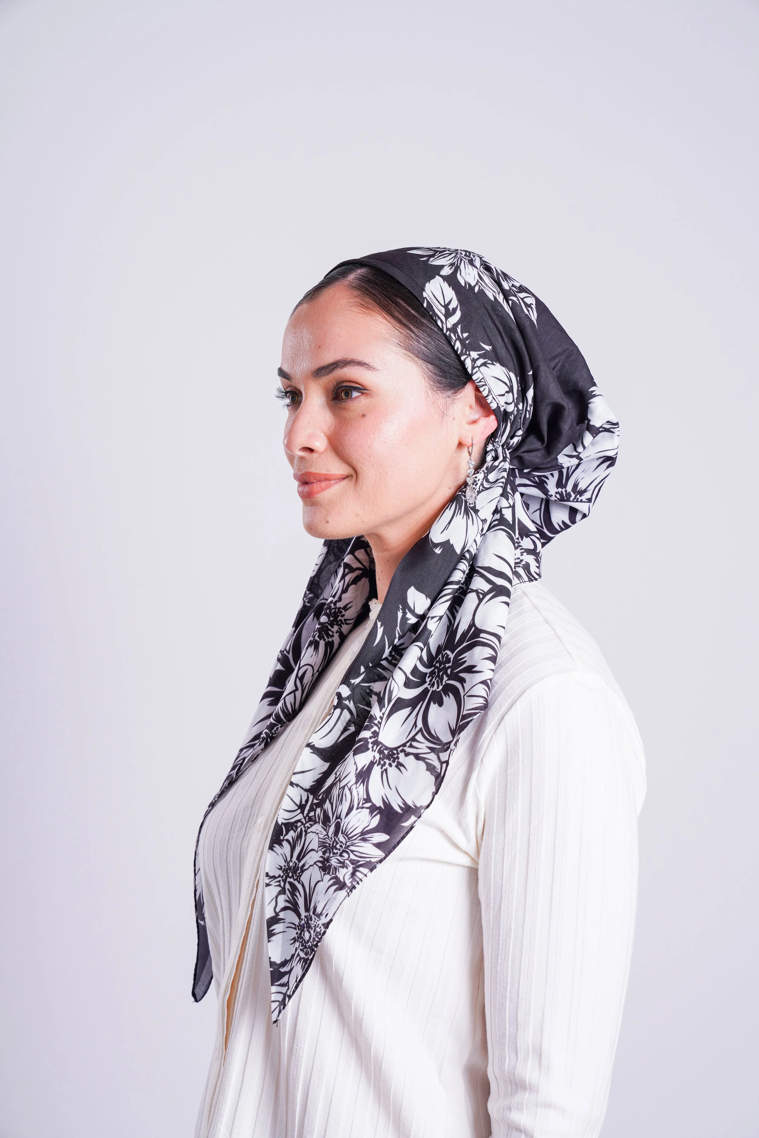 A woman with dark hair wearing a black and white floral headscarf and a cream-colored top, posing against a plain white background.