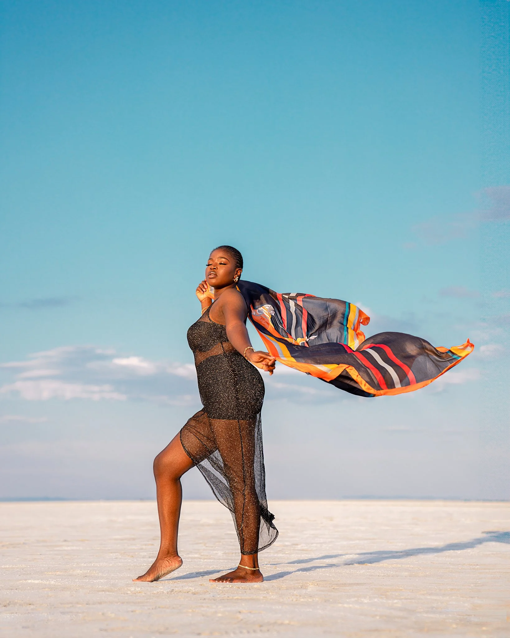 A woman standing barefoot on a sandy beach with her eyes closed, wearing a sheer black dress, holding a colorful scarf that is flowing in the wind, under a clear blue sky.