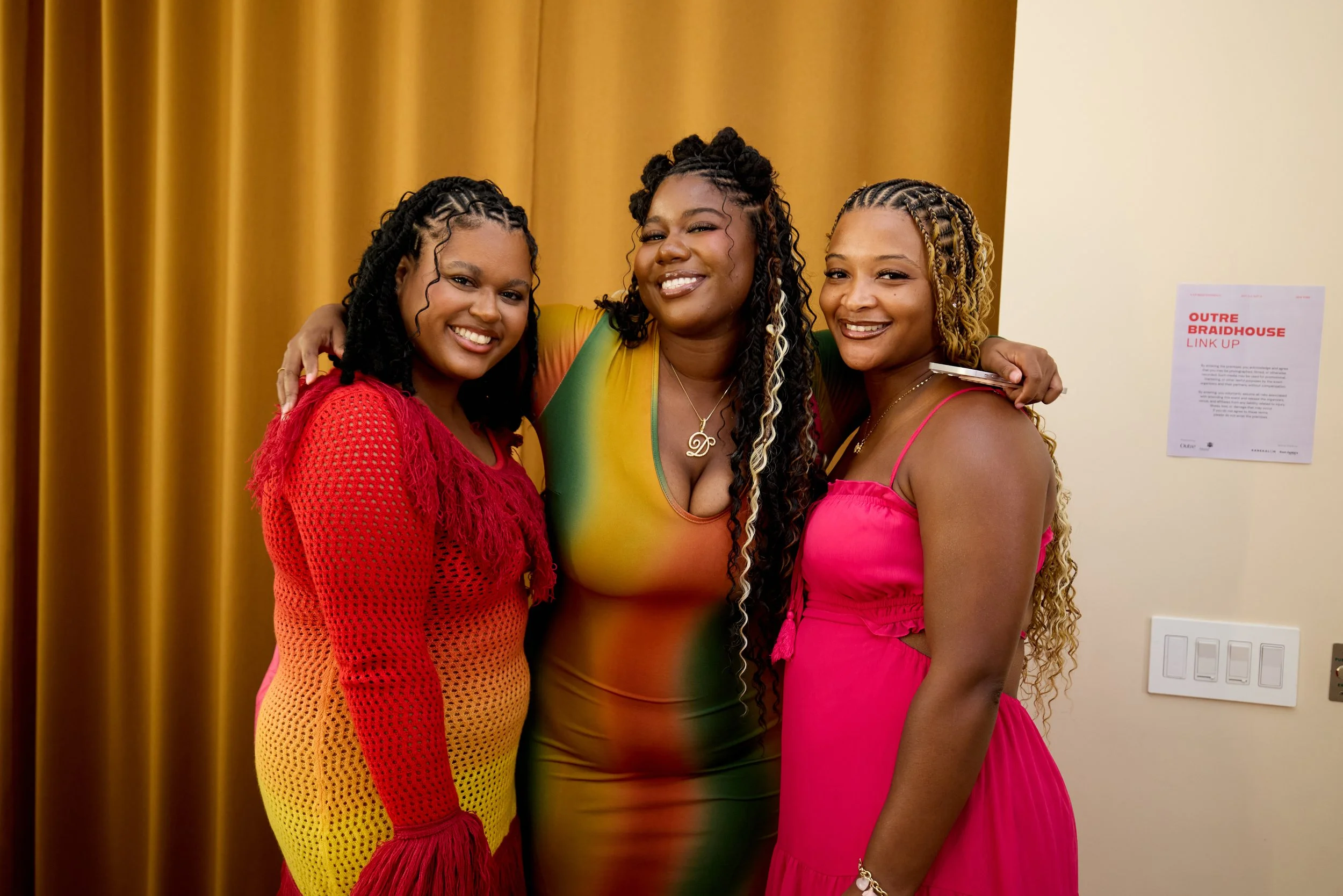 Three women smiling and hugging each other, standing in front of a gold curtain, dressed in colorful dresses.