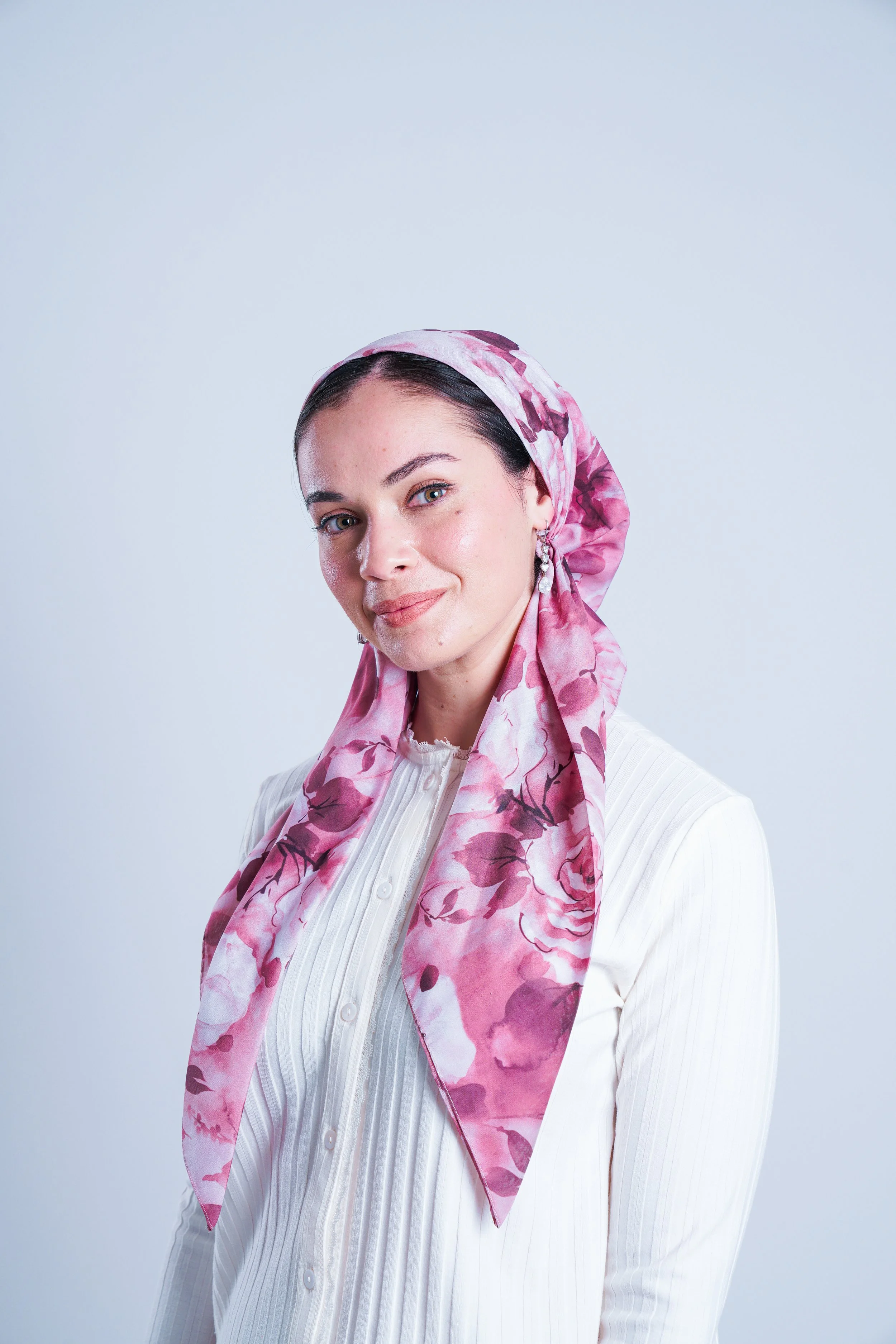 A woman with dark hair, wearing a pink floral scarf and white blouse, smiling softly against a plain light background.