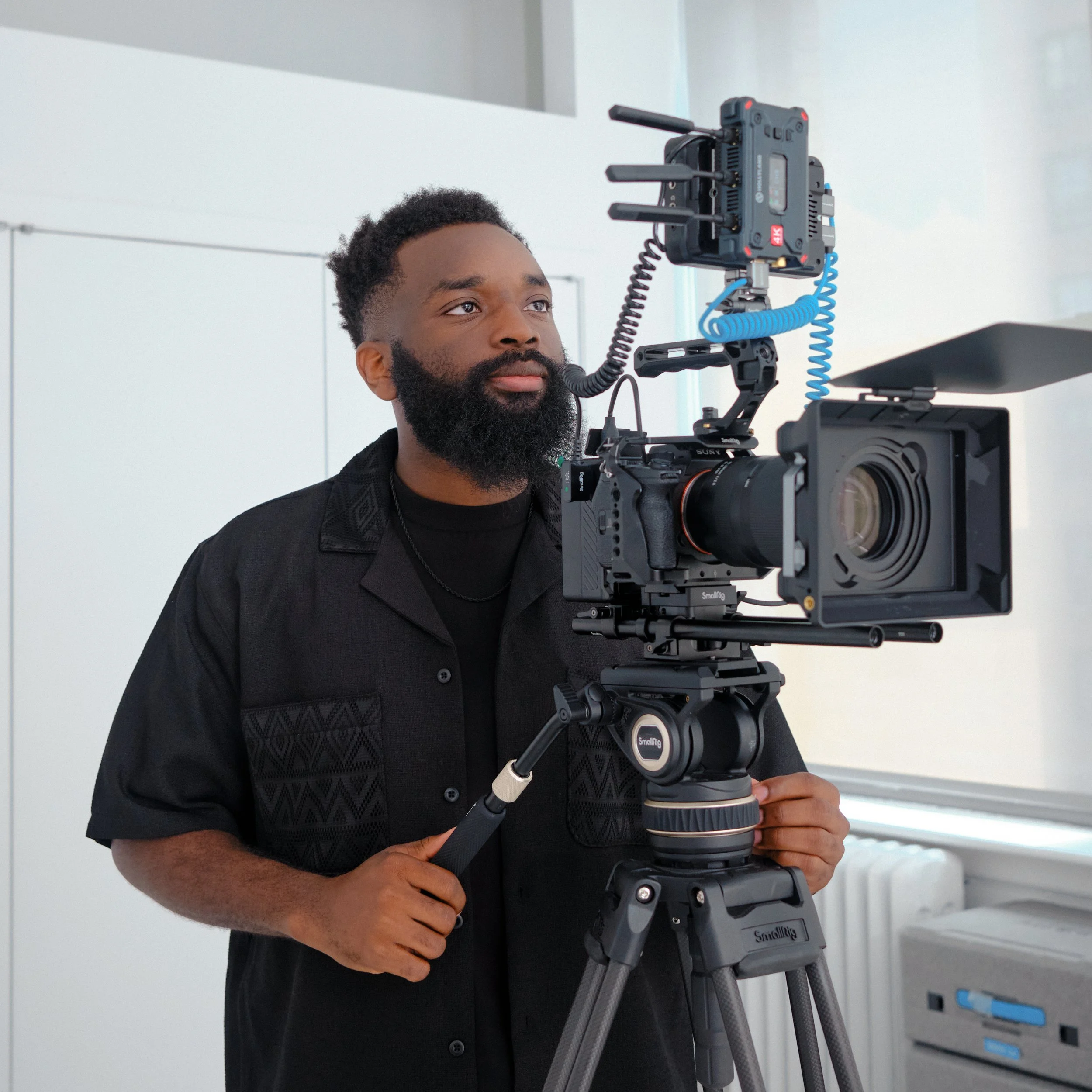 A man with a beard operating a professional camera on a tripod in a well-lit room.