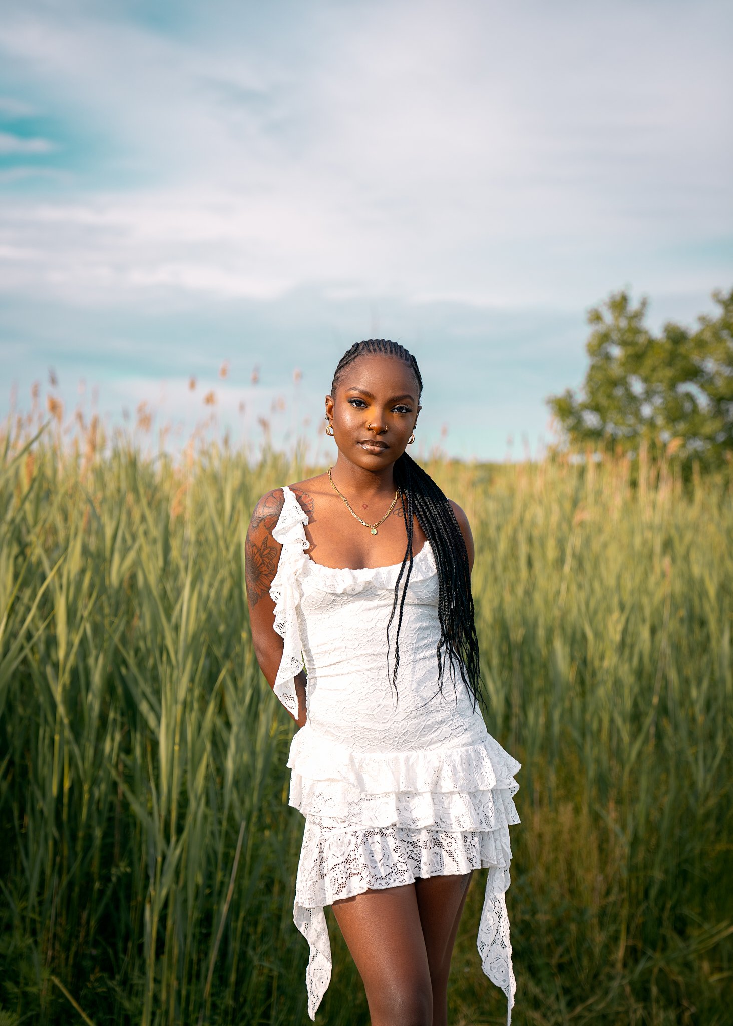 A woman stands in a field of tall grass, wearing a white lace dress with ruffles. She has long braided hair and is looking at the camera with a neutral expression.