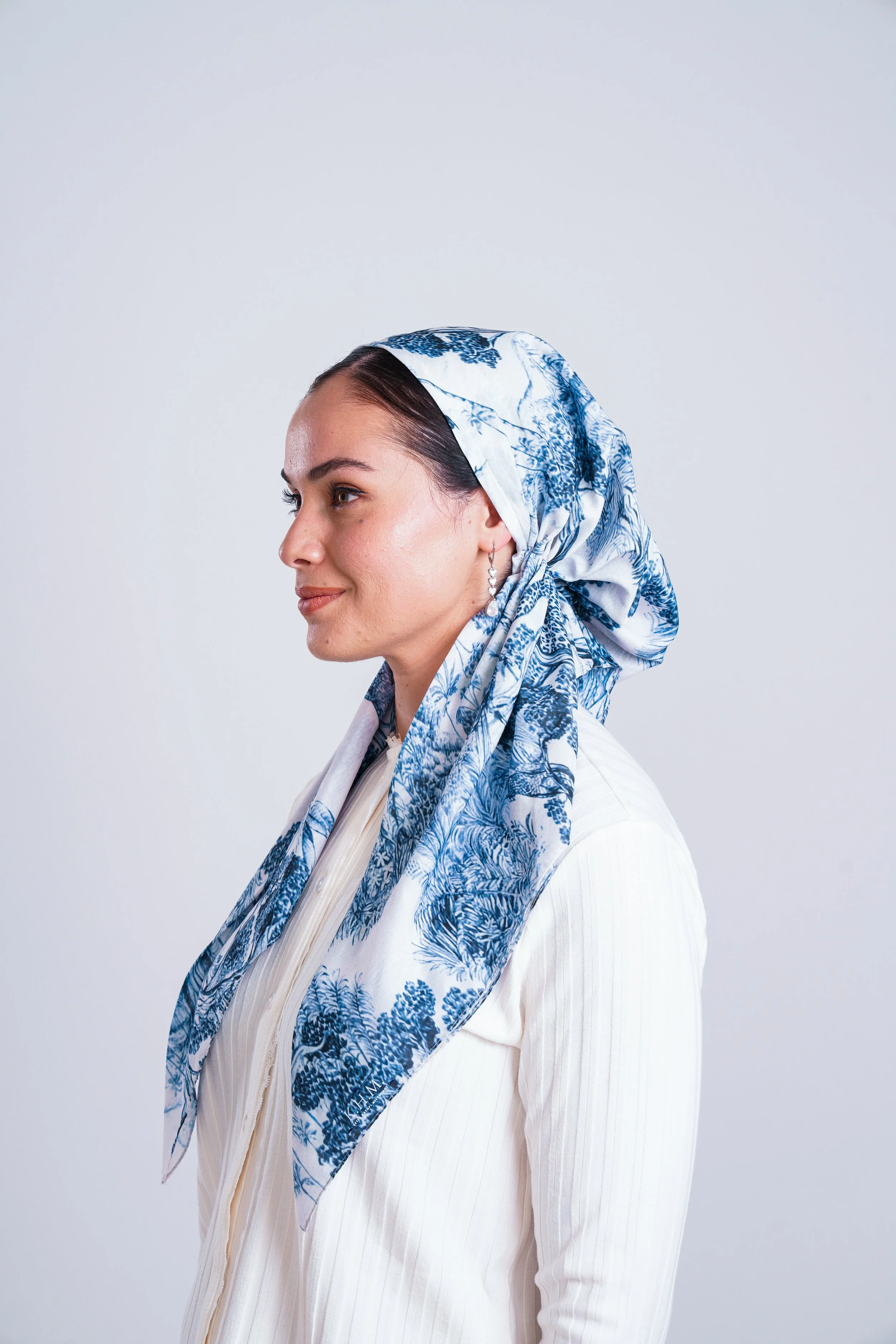 Side profile of a woman with dark hair wearing a blue and white patterned headscarf and a white textured long-sleeve top, standing against a plain light grey background.