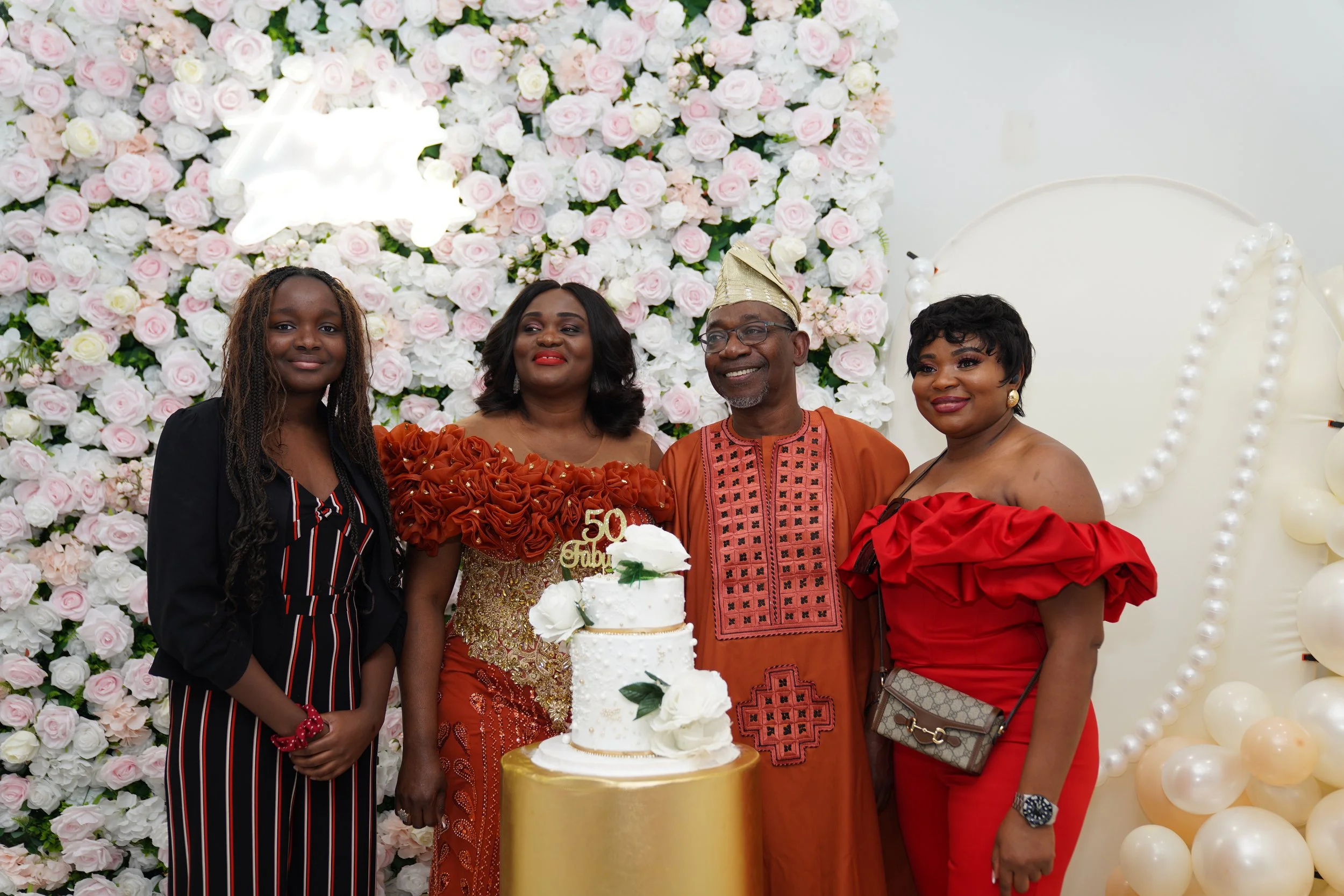Family celebrating a 50th anniversary with a decorated cake, standing in front of a floral backdrop and balloon decorations.
