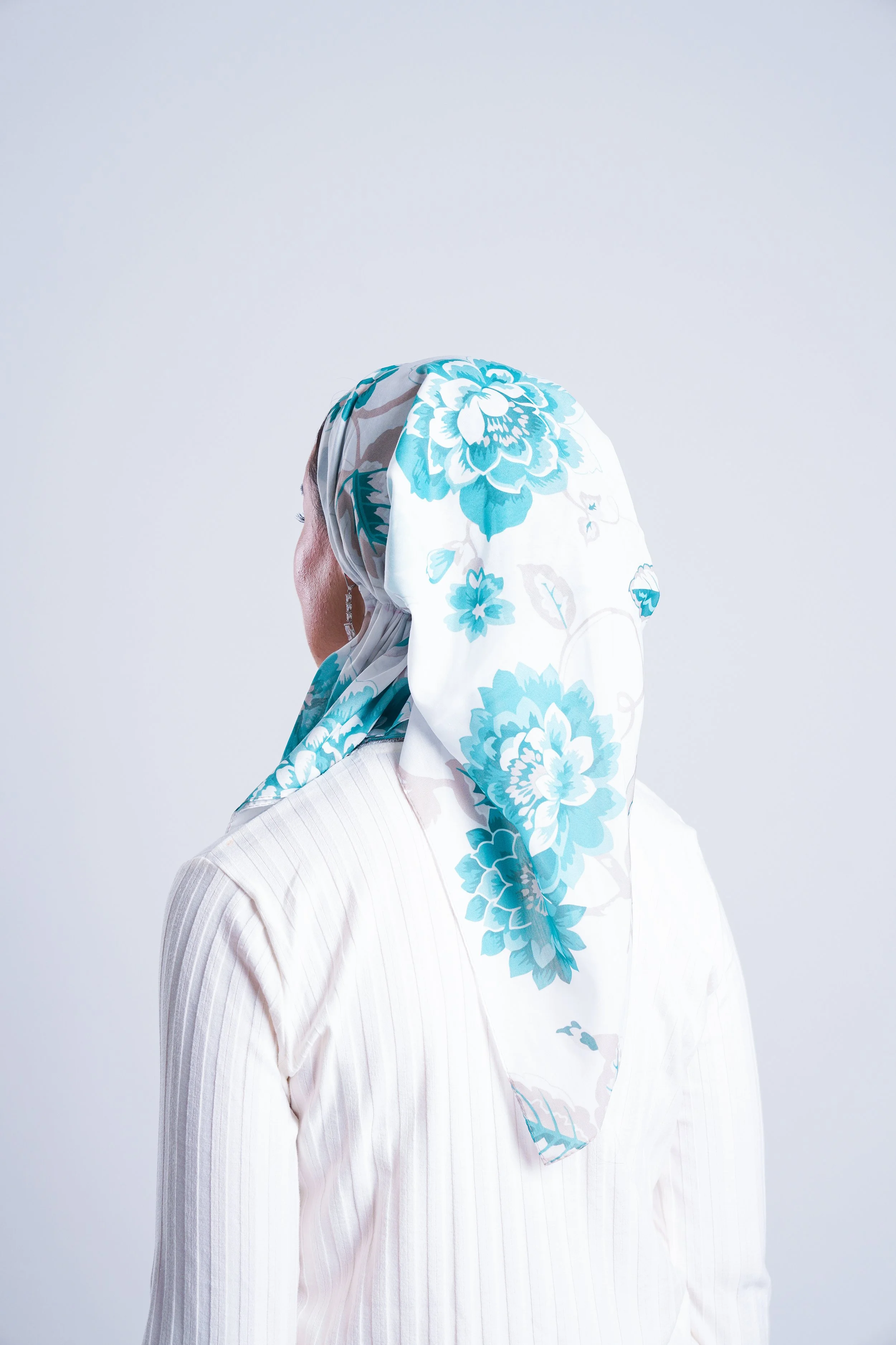 A person wearing a white blouse with vertical stripes and a floral headscarf with blue and white flowers, turned to the side against a plain light background.