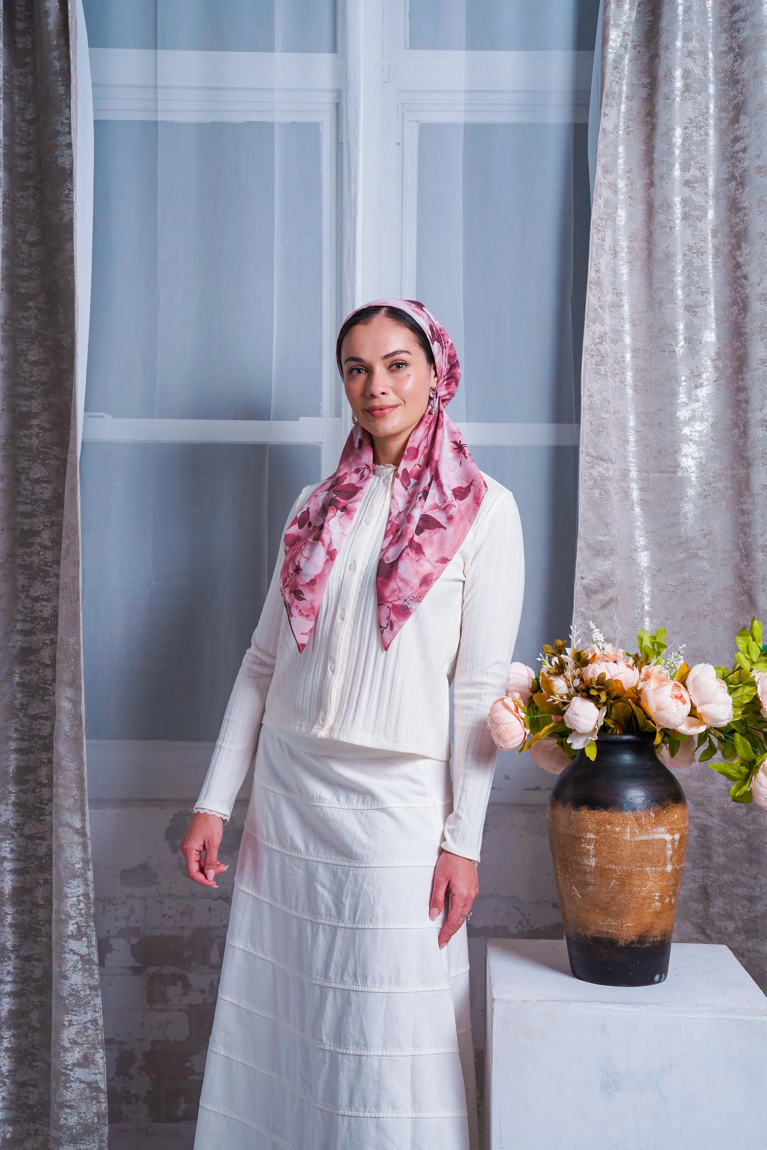 A woman standing indoors in front of large window with light blue curtains. She is wearing a white cardigan and skirt with a pink patterned headscarf, and has a vase with pink and white flowers beside her.