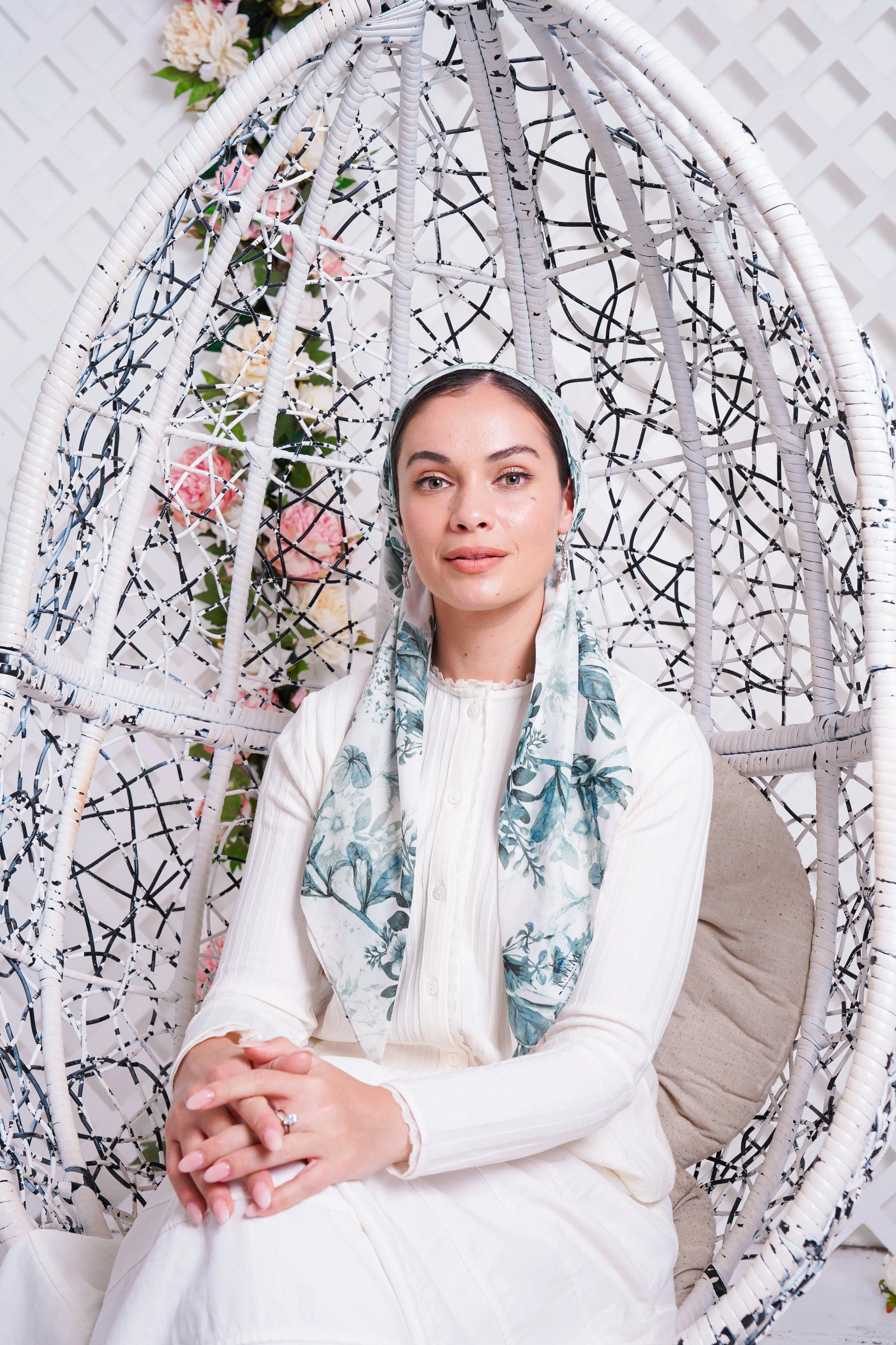 A woman sitting in a white wicker hanging chair with a floral pillow, wearing a white dress and a light-colored scarf with blue and green leaf patterns, in front of a backdrop decorated with pink and white flowers.
