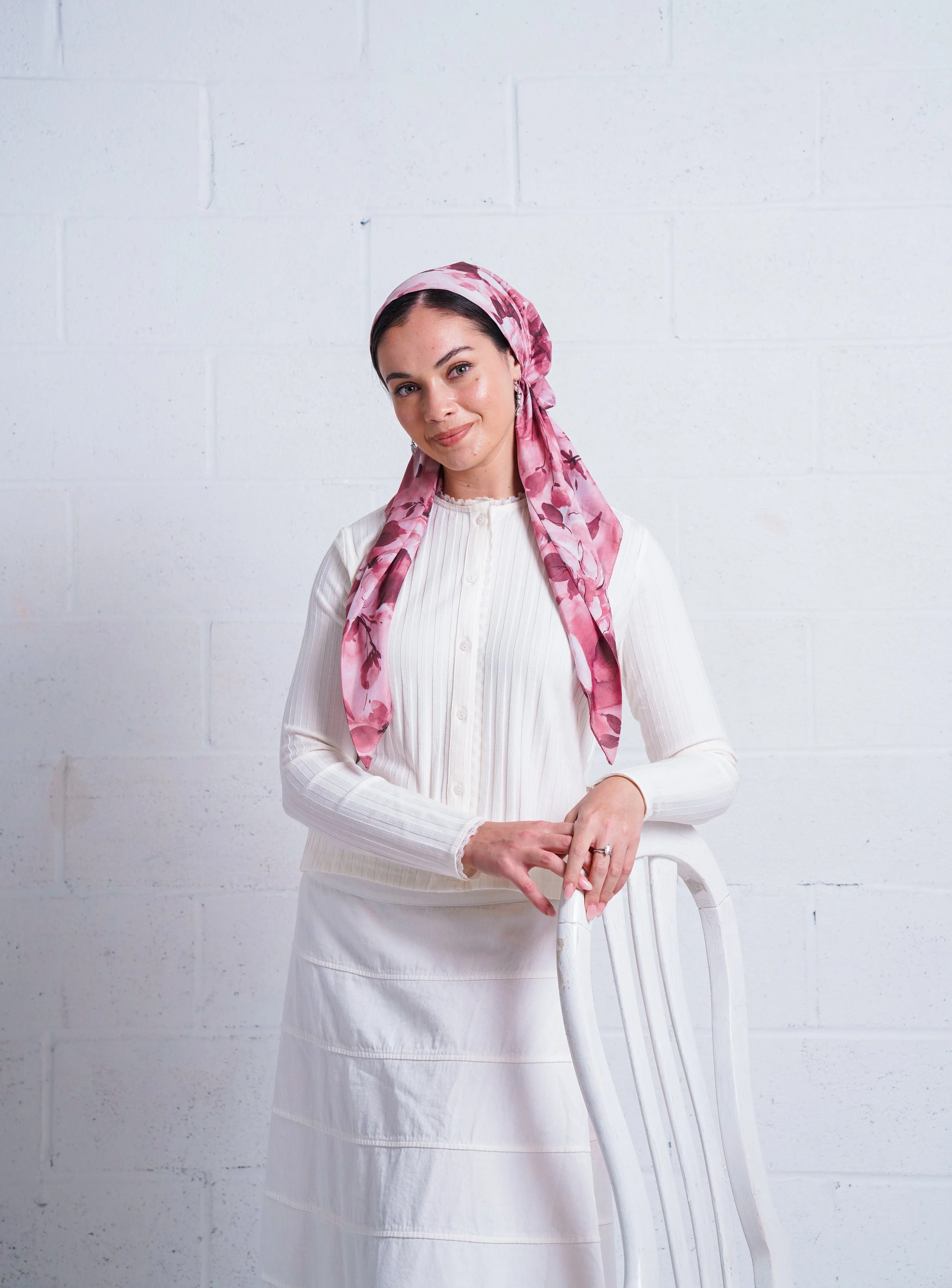 A woman with dark hair and light skin, wearing a pink floral headscarf, a white long-sleeved shirt, and a white skirt, leaning on a white chair against a plain white wall.