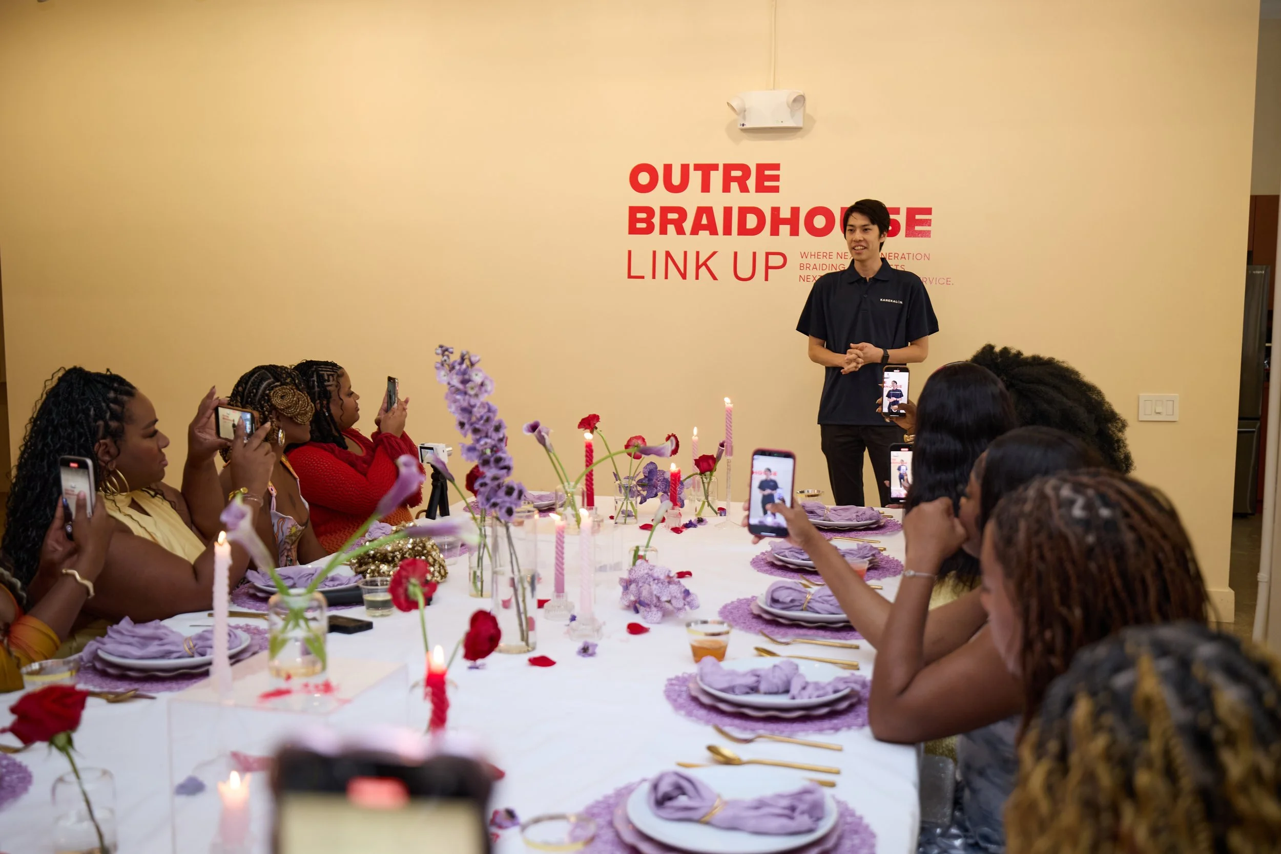A woman speaker stands in front of a group of women at a table, giving a talk or presentation at an event with purple and red floral decorations, candles, and purple table settings. The background has a beige wall with red text that reads 'OUTRE BRAI