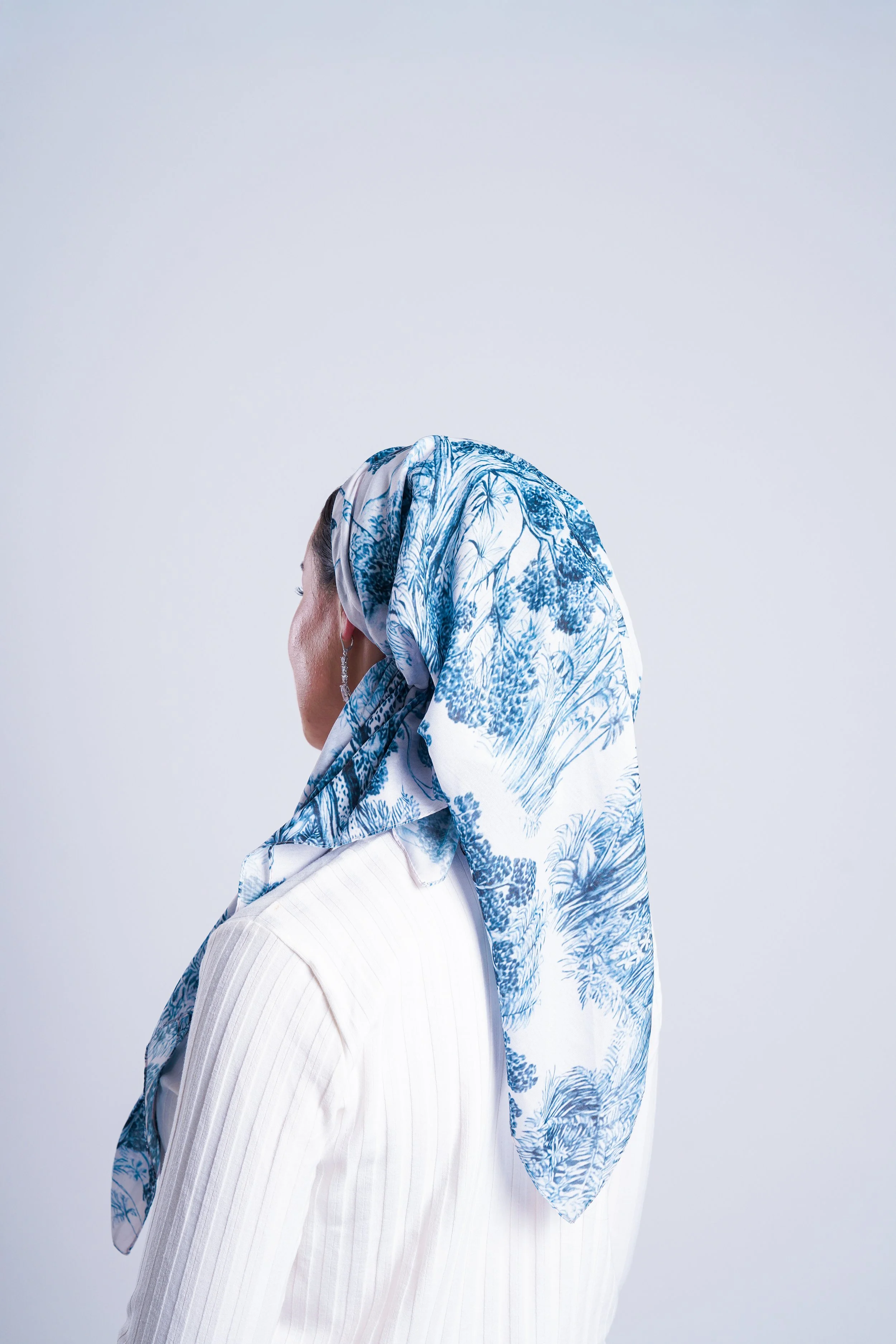 A woman wearing a white blazer and a blue and white patterned hijab, seen from the side against a plain white background.