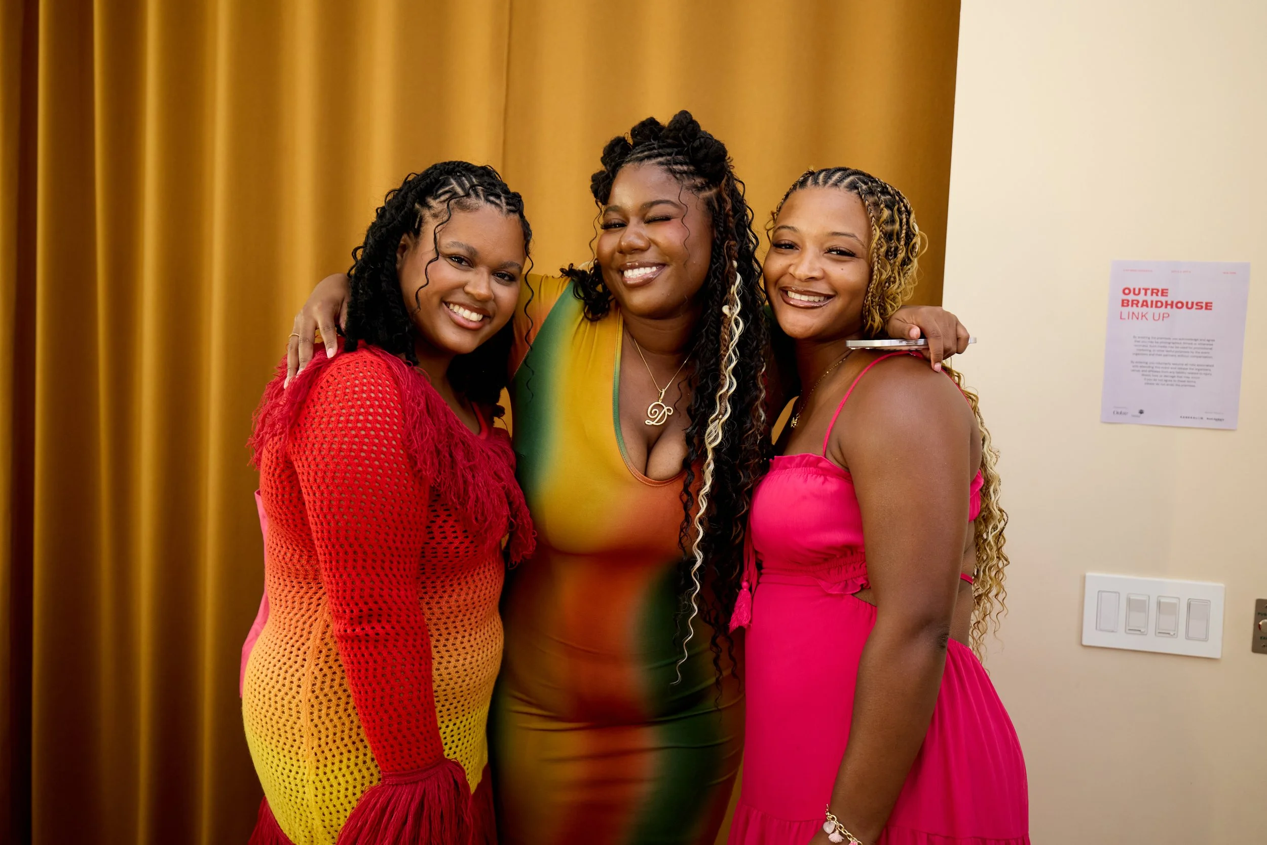 Three women smiling and hugging in front of a gold curtain, wearing colorful dresses for a festive event.