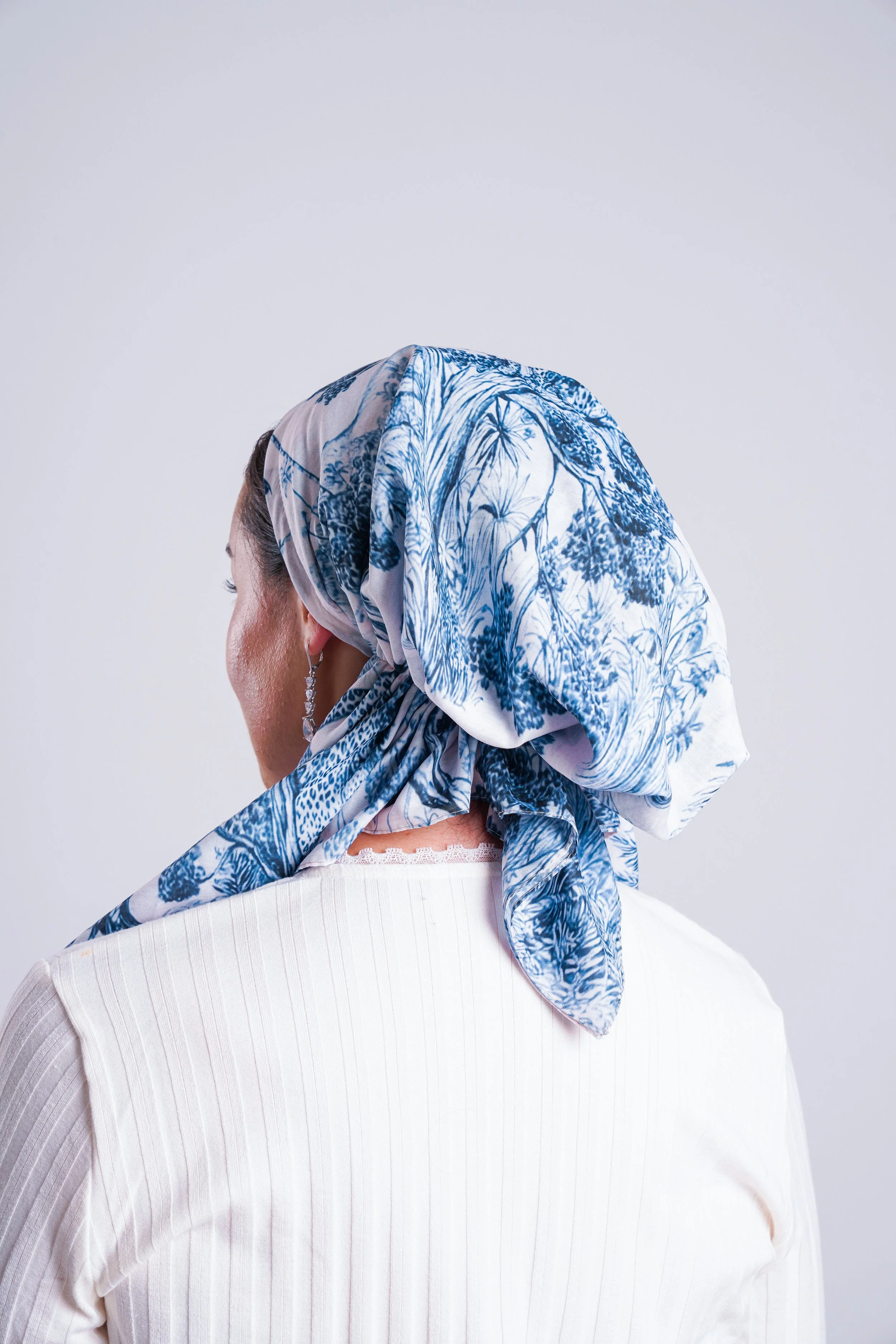 A woman wearing a white and blue patterned headscarf and a white top, facing away from the camera with a plain light-colored background.