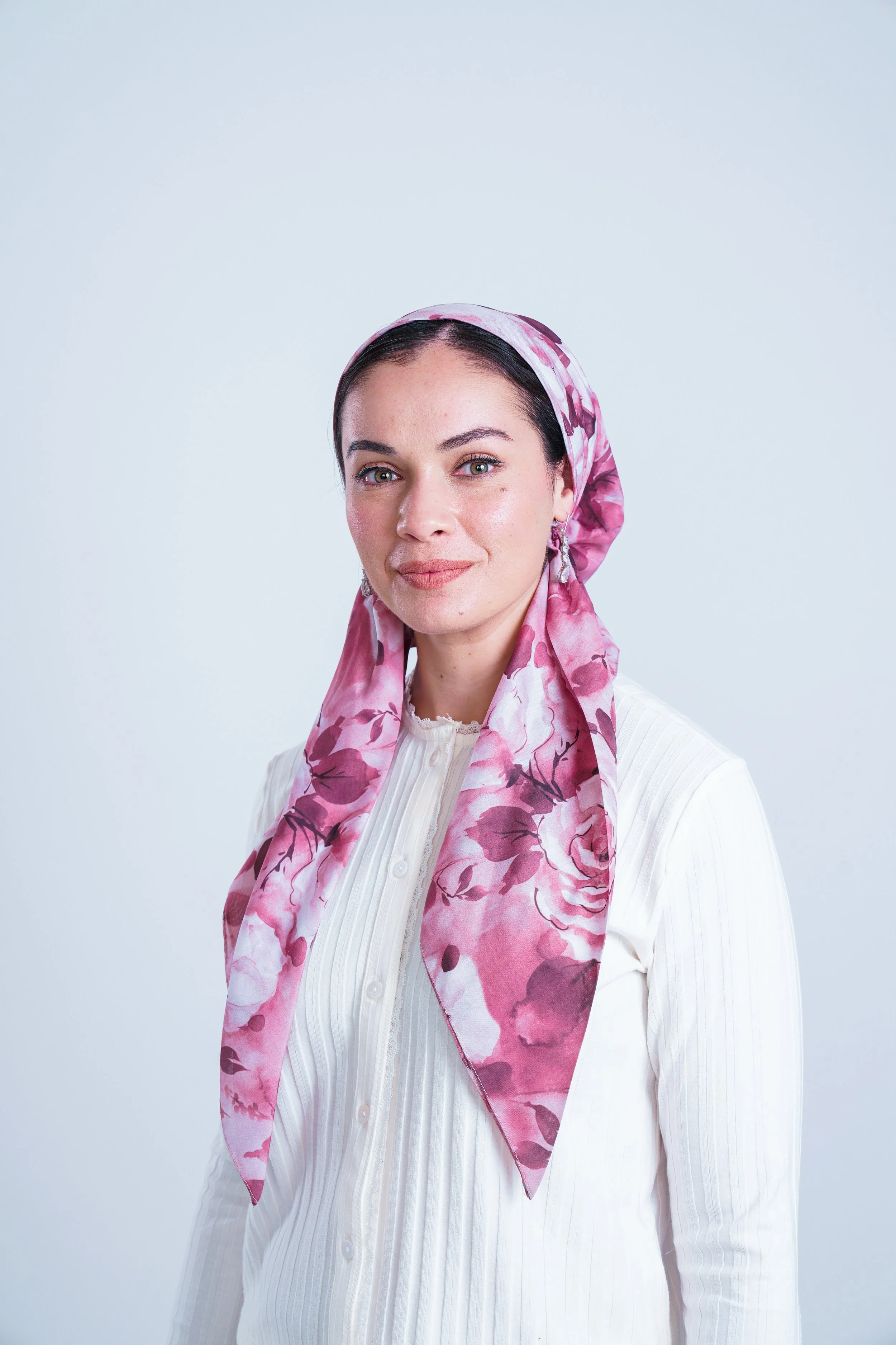 Portrait of a woman wearing a pink floral headscarf and a white blouse, smiling lightly against a plain background.