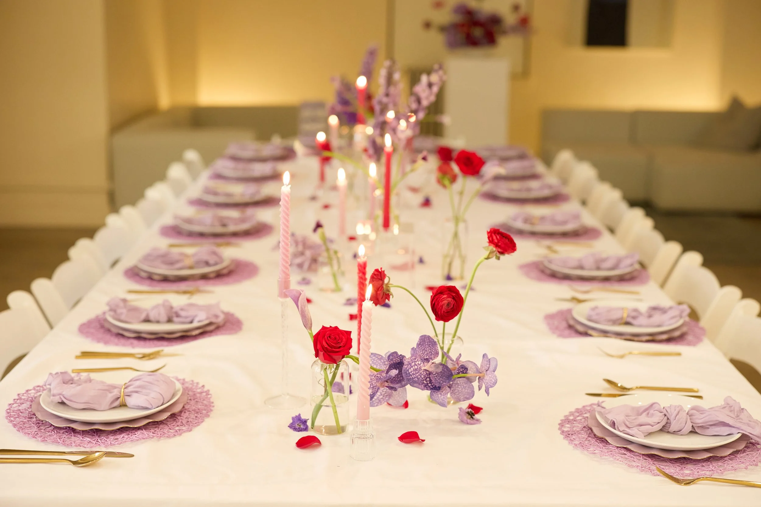 A long dining table decorated with pink candles, red and purple flowers, pink napkins, gold cutlery, set for a celebration in a warmly lit room.