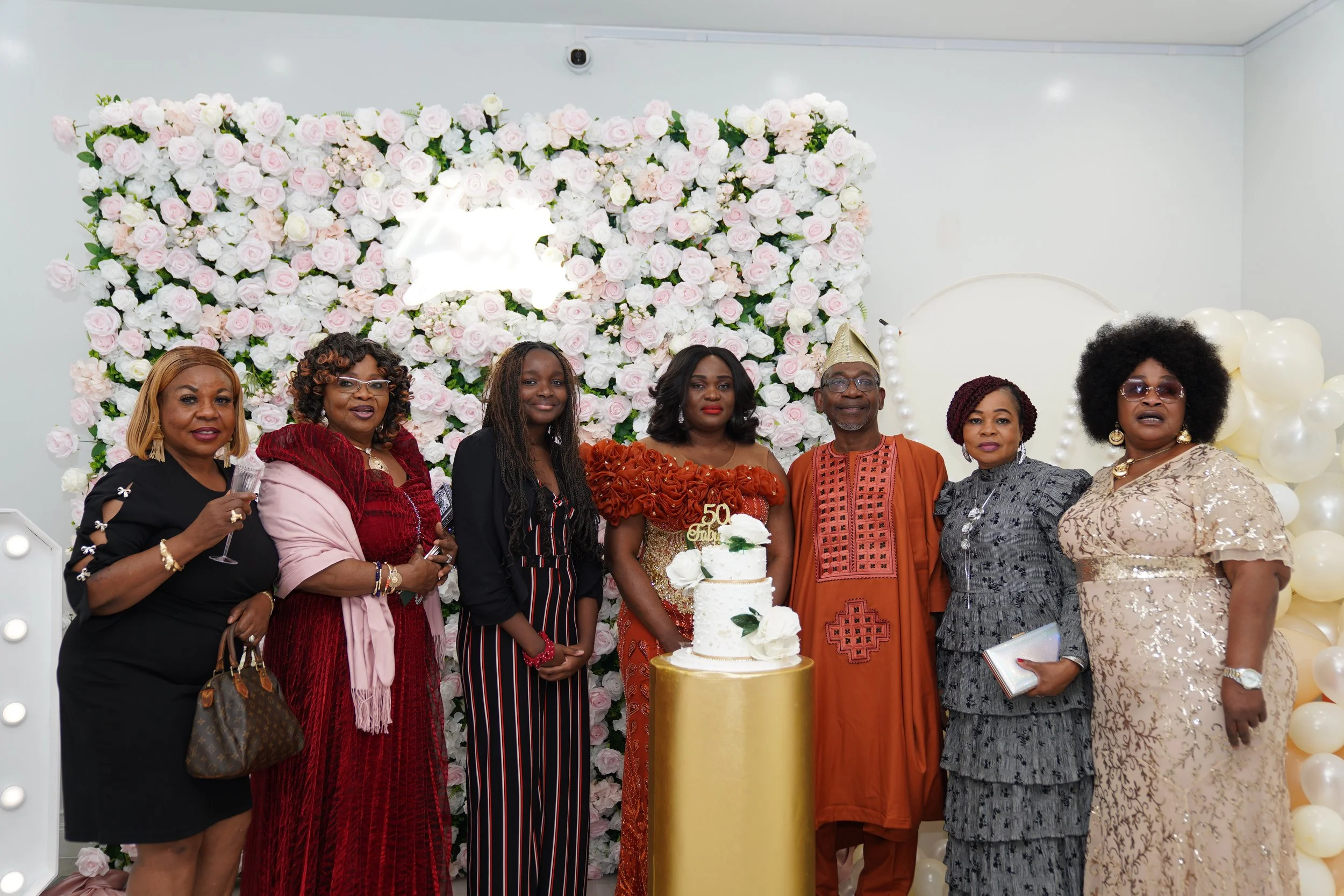 Group of people celebrating a 50th birthday with a cake, standing in front of a floral backdrop with balloons.