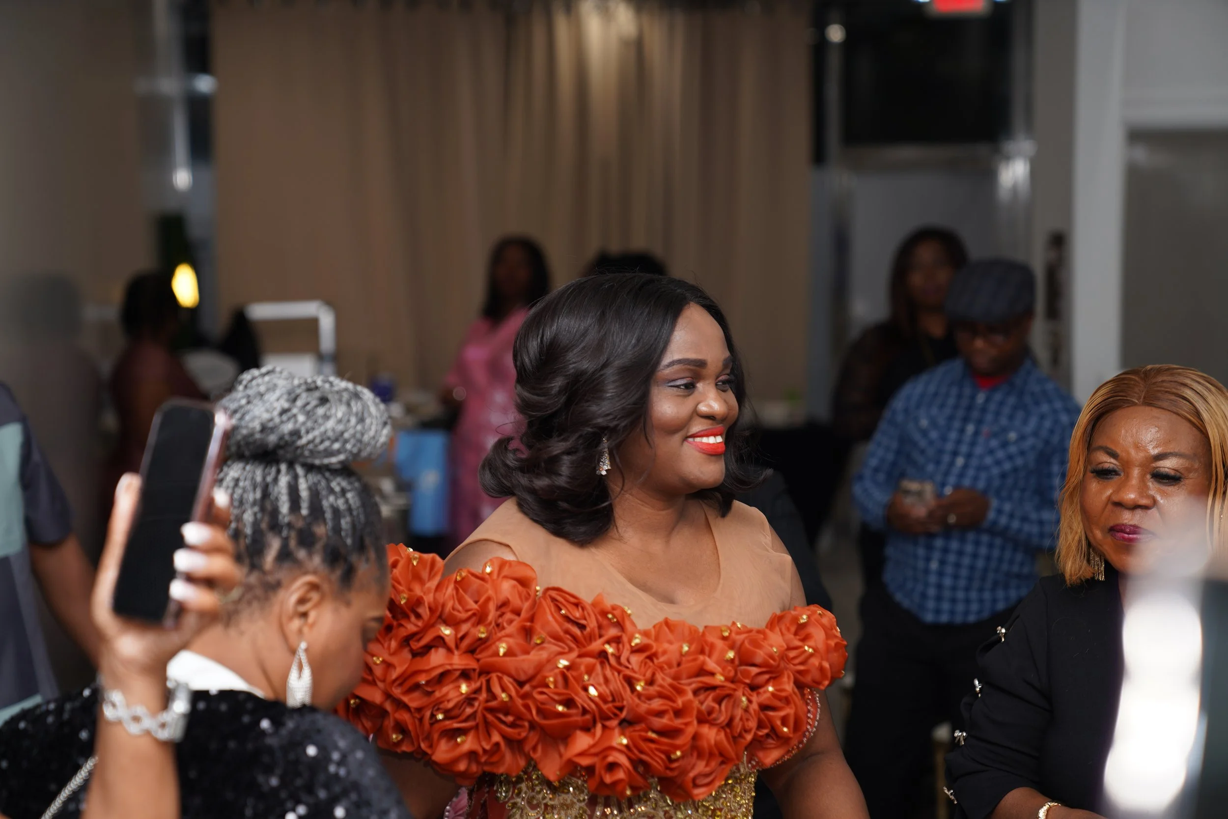 A woman in a beige dress with orange floral ruffles on the shoulders, smiling at an event with people in the background.
