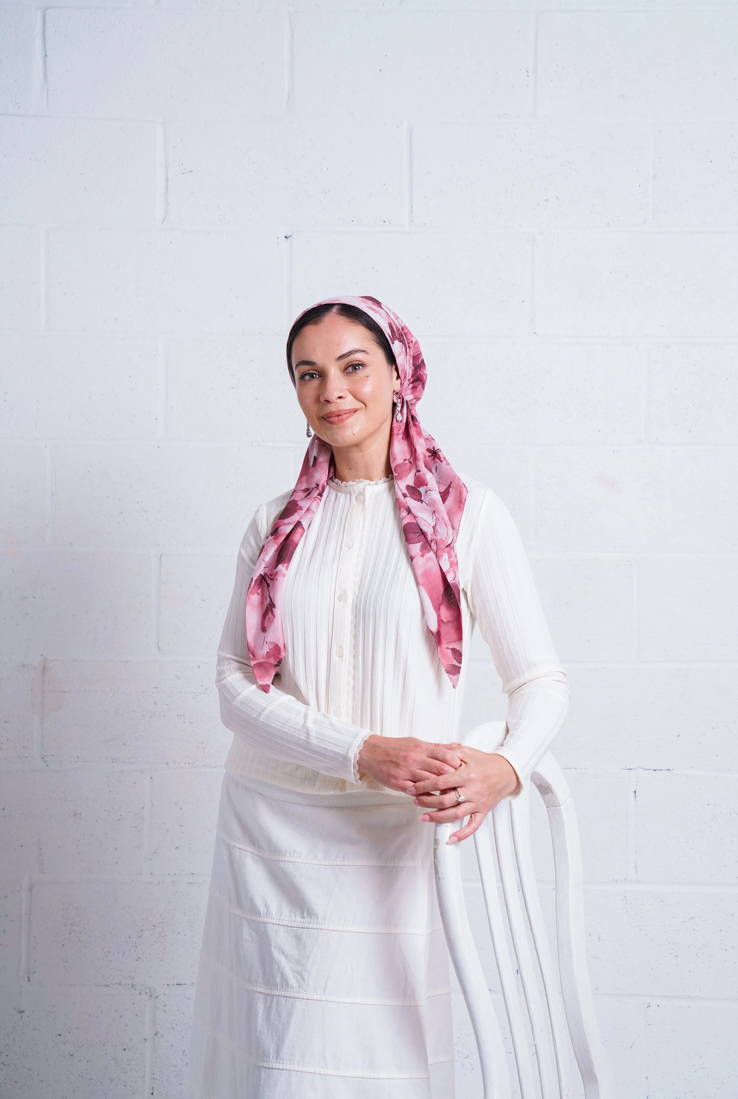 A woman with dark hair and fair skin wearing a pink floral headscarf, a white long-sleeve blouse, and a white skirt standing against a white brick wall, holding a white chair.