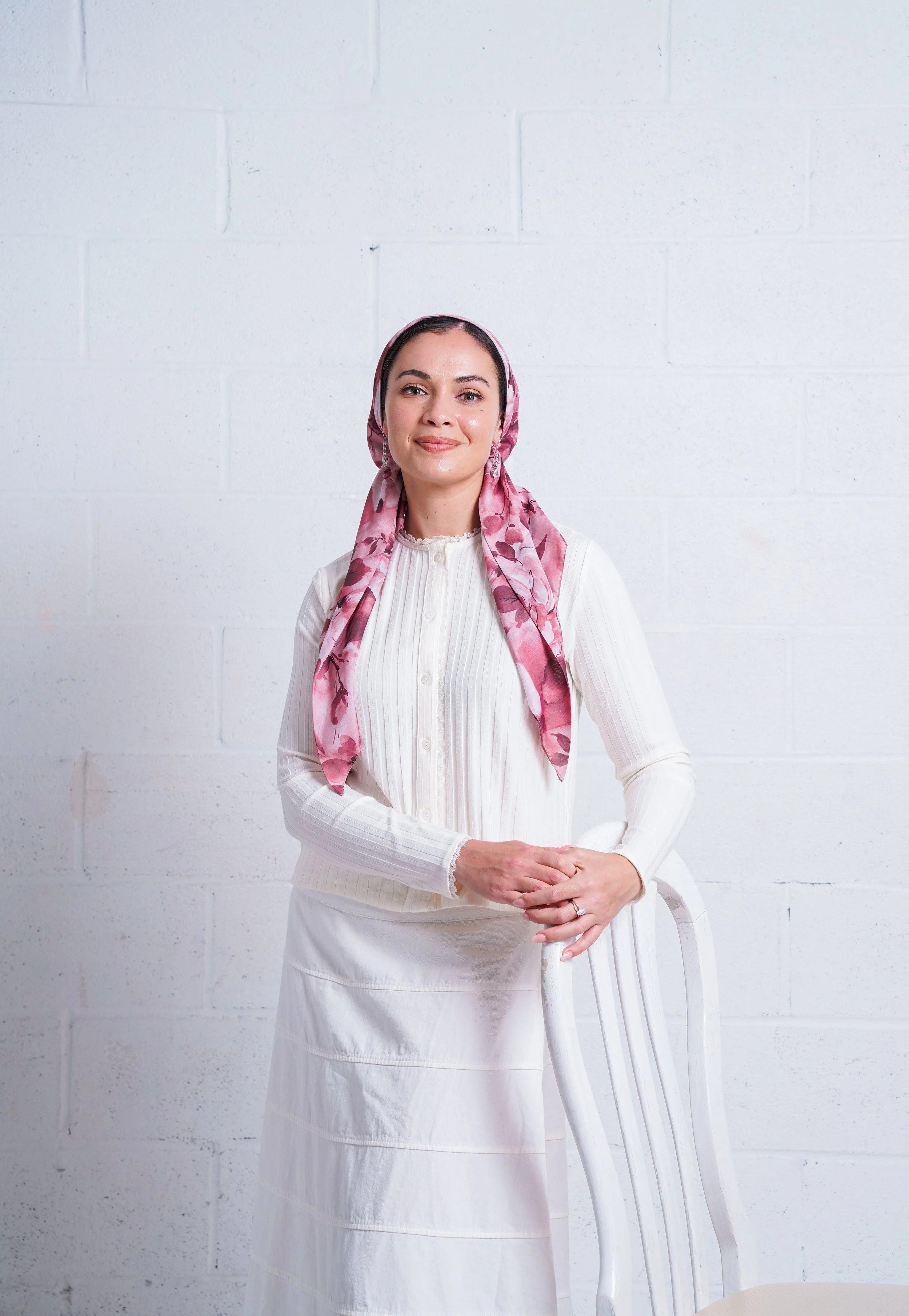 Woman wearing a white outfit and pink floral headscarf standing against a white brick wall, smiling and leaning on a white chair.