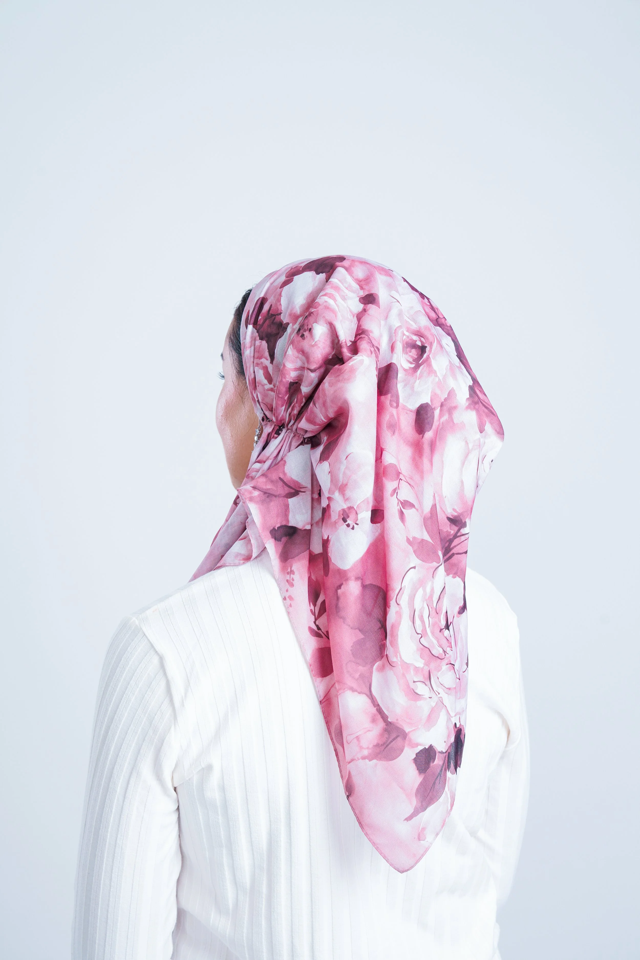 A woman in a white shirt wearing a pink floral headscarf, facing to the left against a plain light background.
