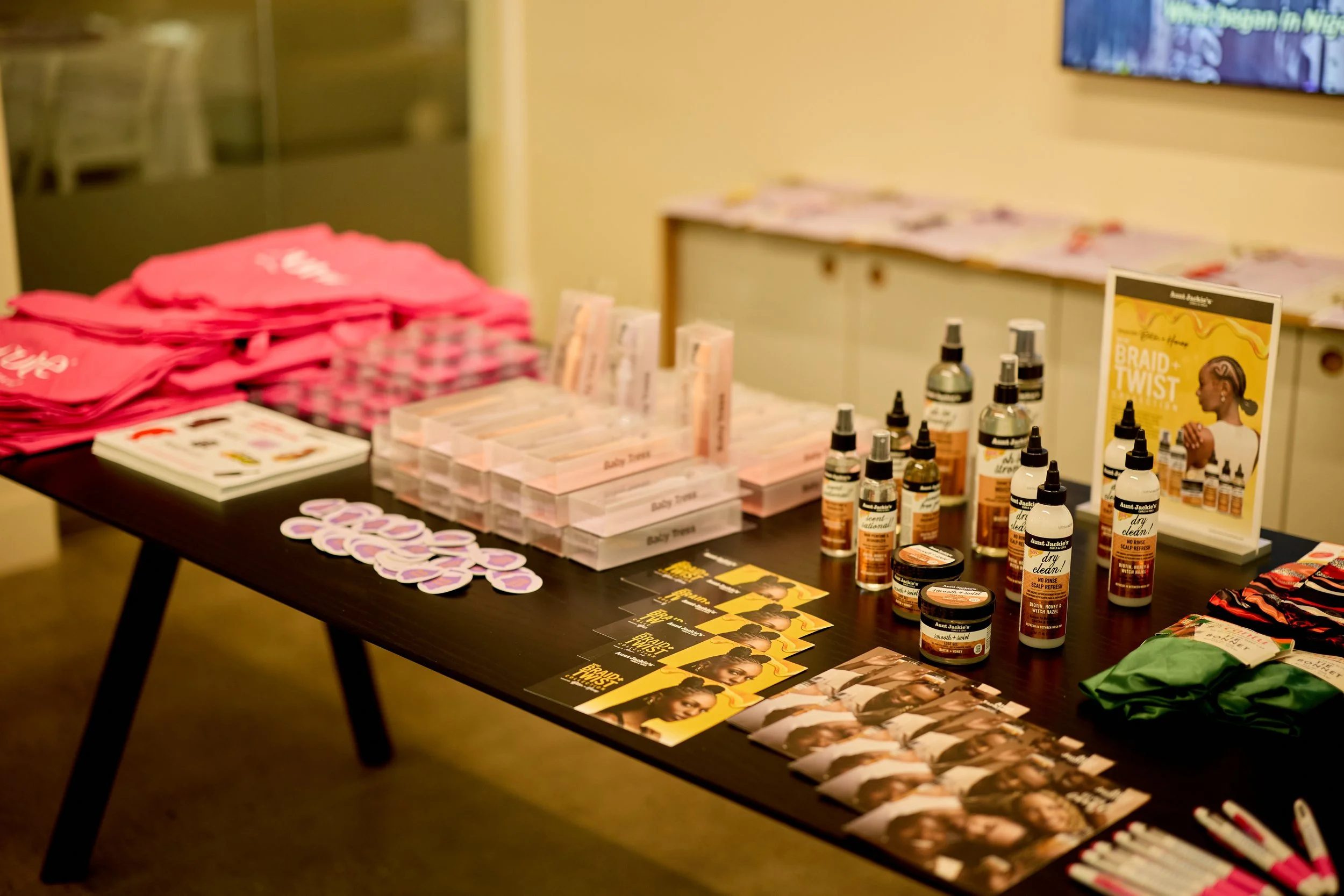 A black table displaying hair care products, pink tote bags, stickers, brochures, and promotional materials at an event or store.