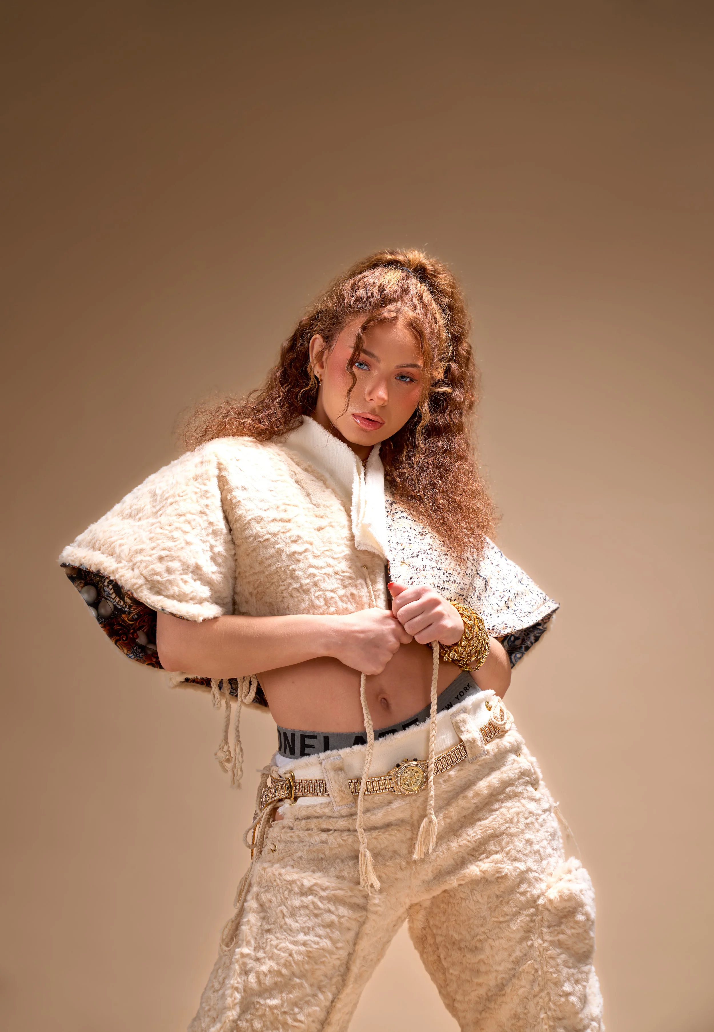 Young woman with curly brown hair wearing beige and white textured loungewear, holding the zipper of her top, standing against a neutral background.
