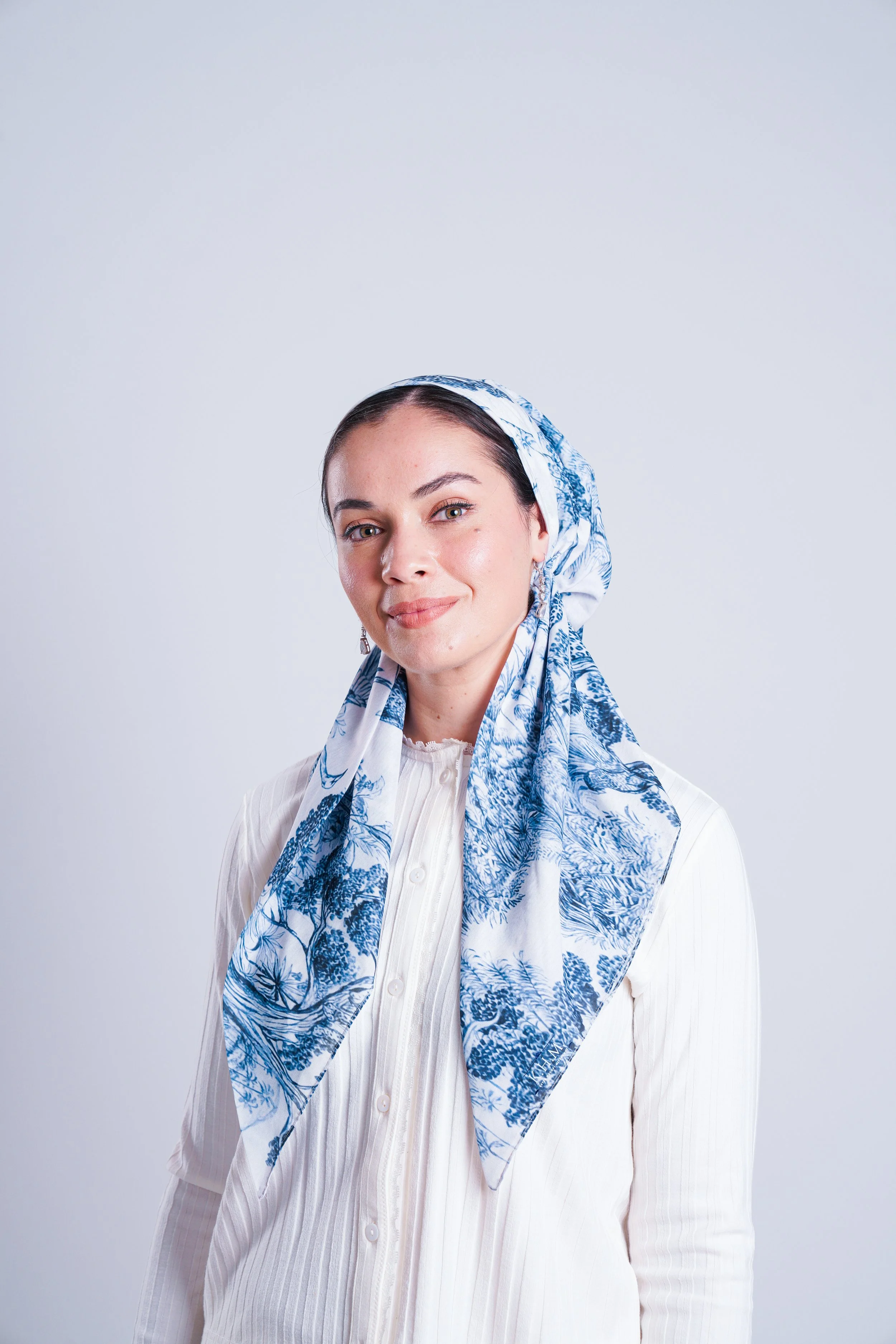 A woman wearing a white blouse and a blue and white patterned headscarf against a plain light gray background.