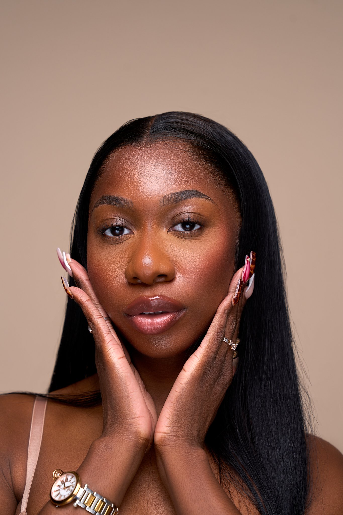 A woman with long black hair and makeup posing with her hands on her face, wearing a watch and rings, against a beige background.