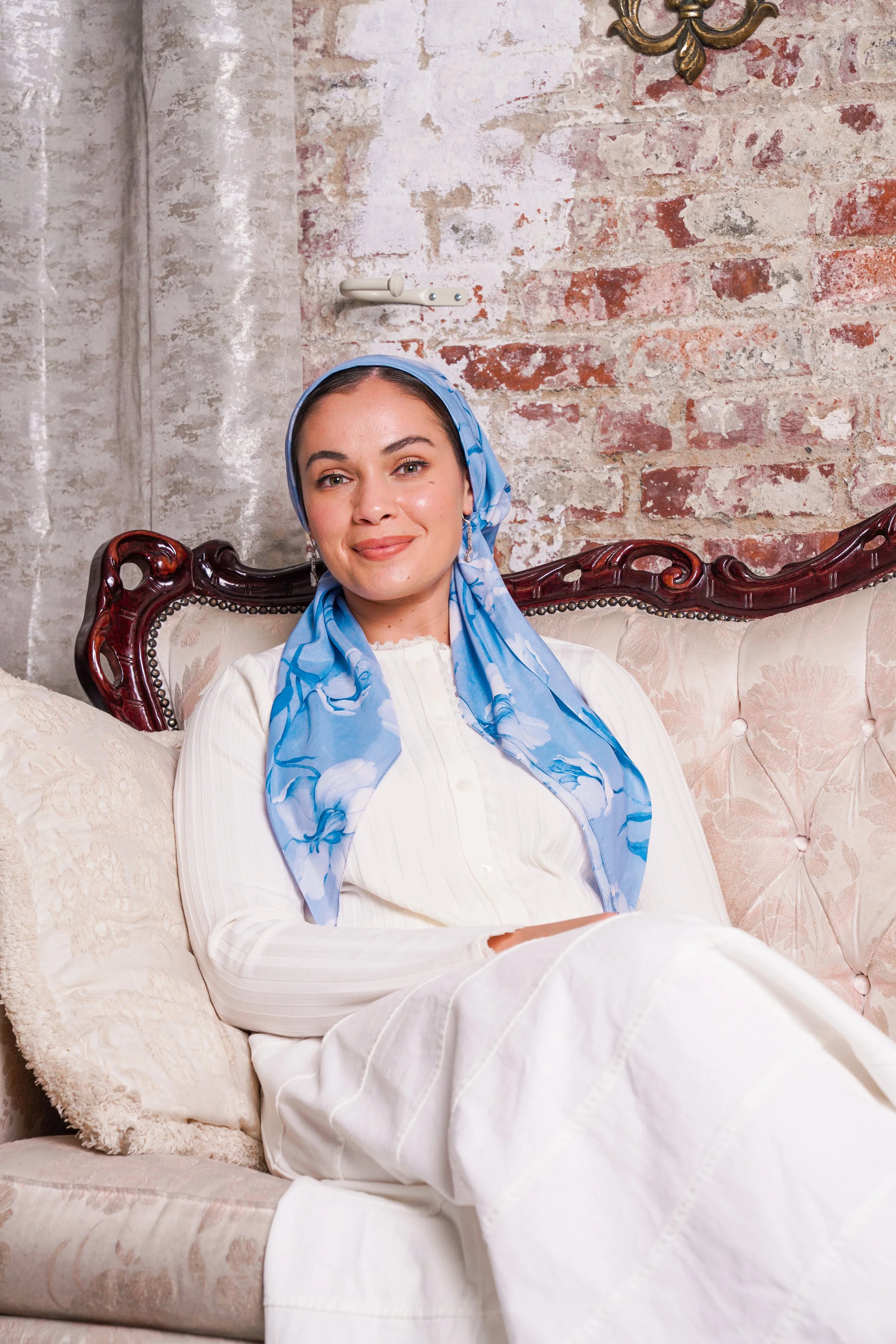 A woman sitting on a vintage sofa with a floral pattern, wearing a white outfit and a blue scarf, smiling at the camera, with a brick wall and an antique wall ornament in the background.