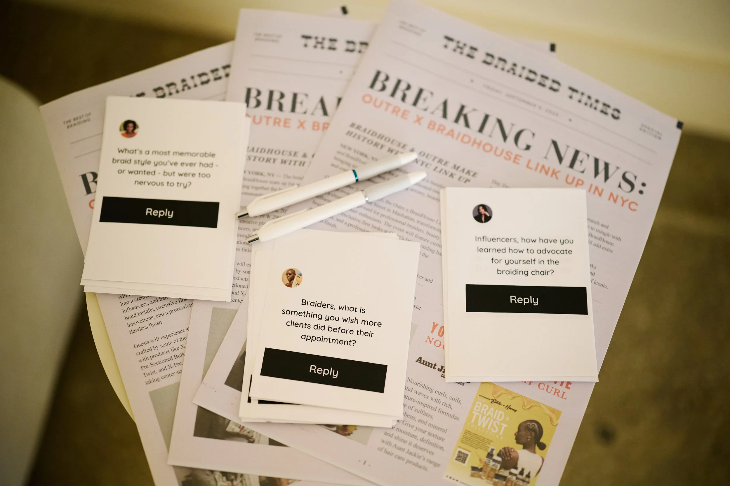 Printed newspapers, pens, and answer cards discussing hair braiding topics on a round table.