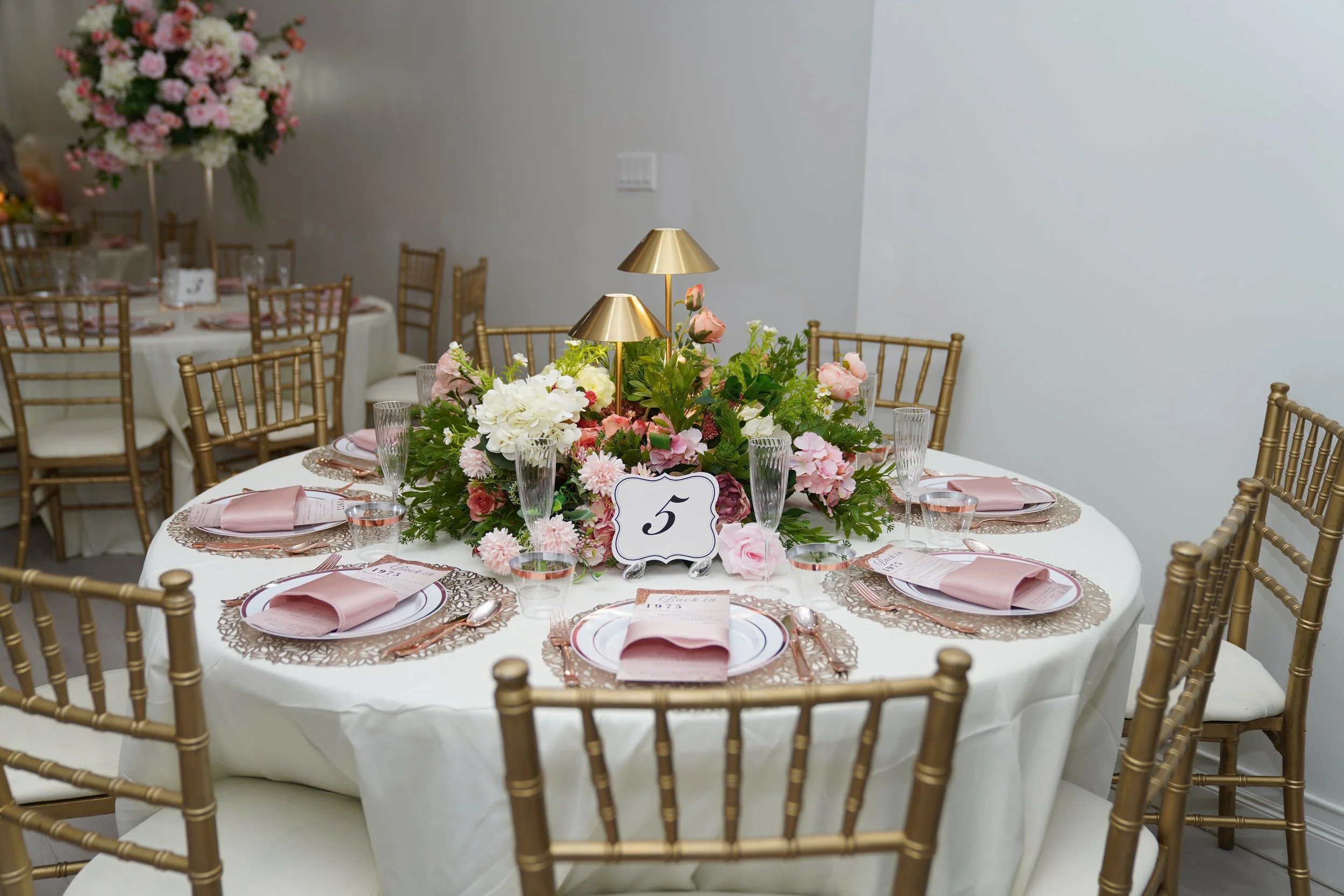 Round banquet table decorated with a floral centerpiece, pink napkins, and gold chairs, set for a formal event with the table number 5.
