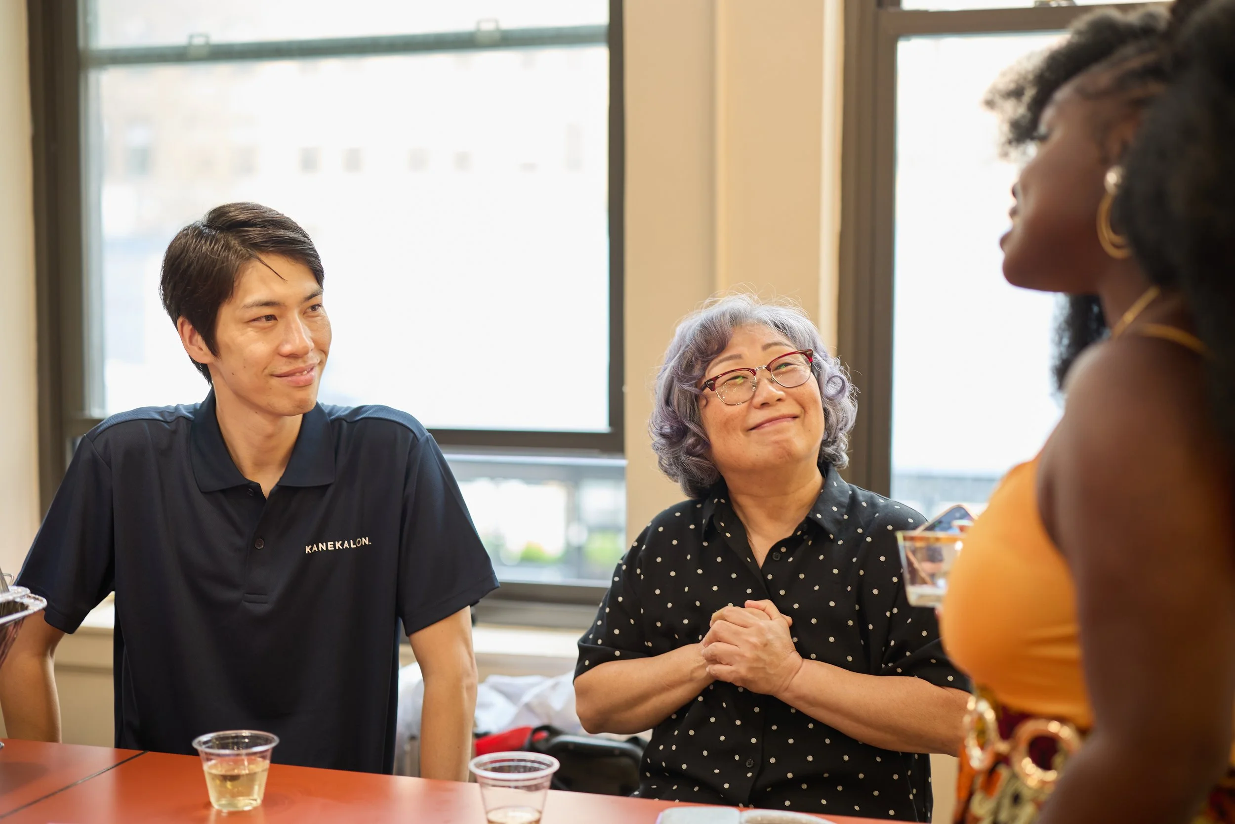 A group of three people, two women and one man, are gathered around a table in a brightly lit room with large windows. They are smiling and engaging in a friendly conversation, each with a drink in front of them.