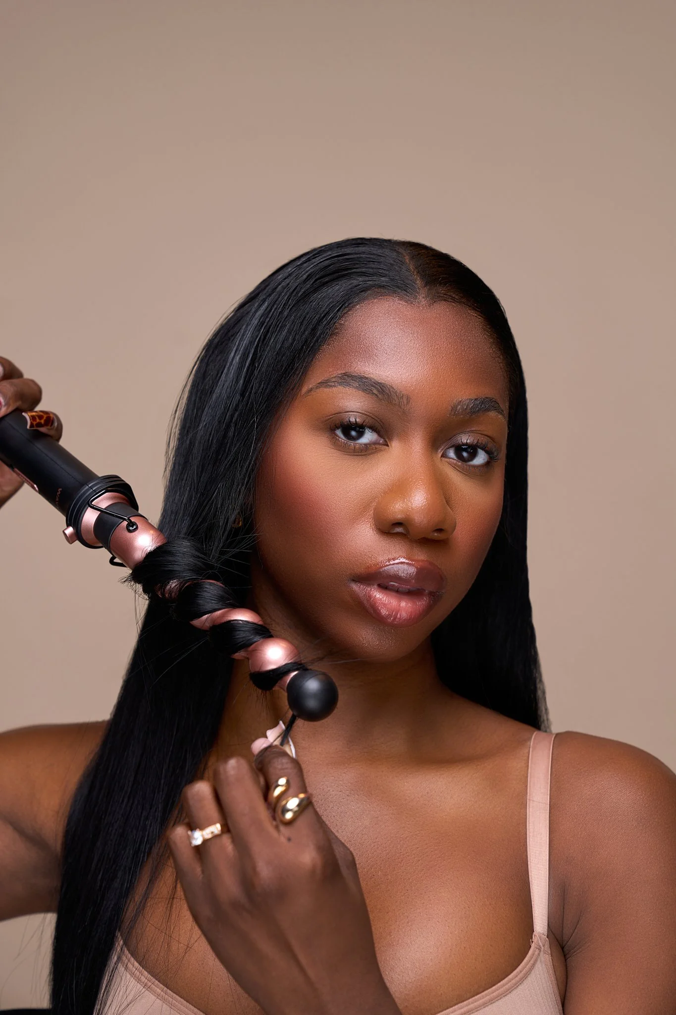 A woman with long black hair using a curling iron on her hair, looking at the camera, beige background.