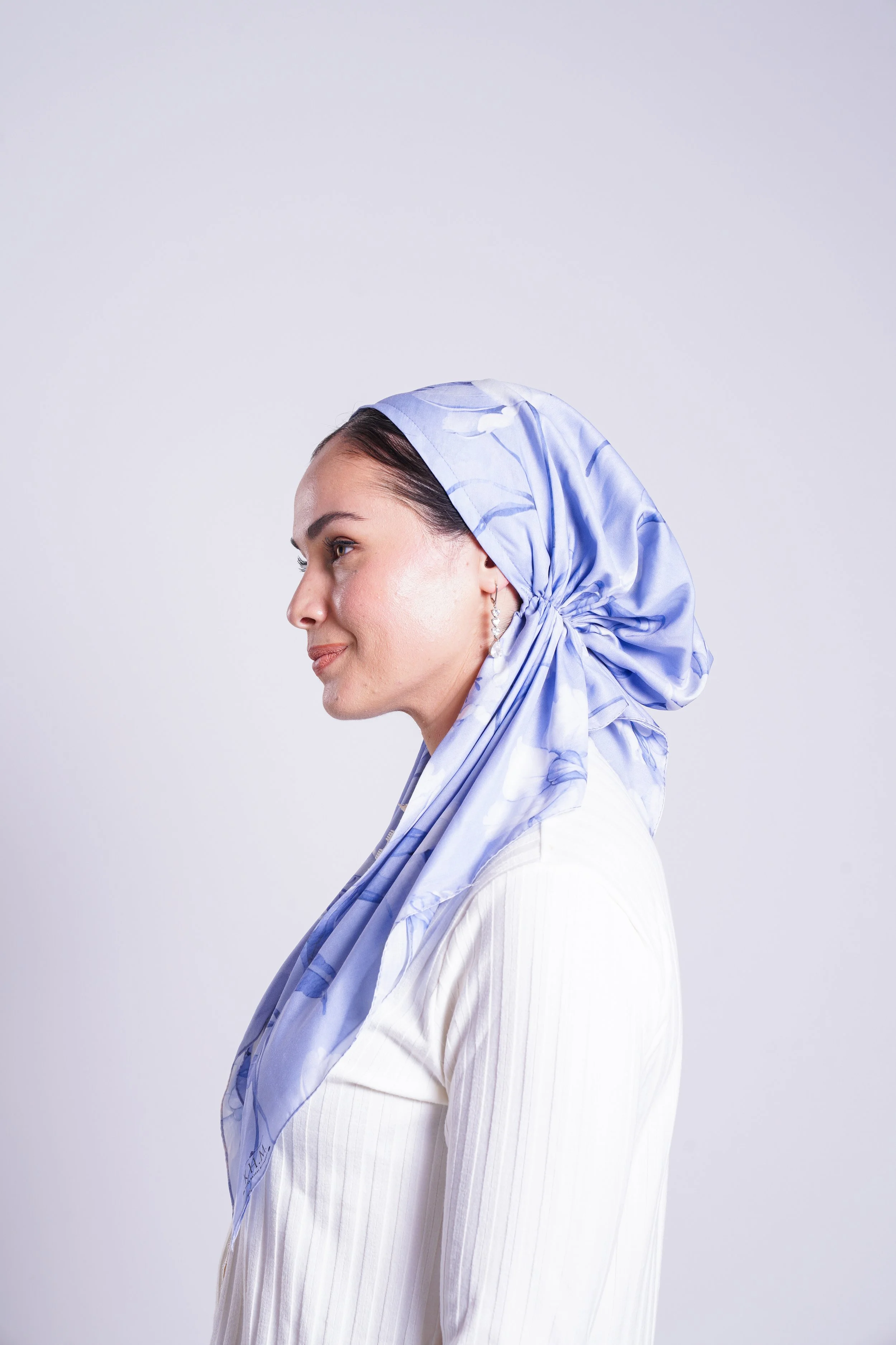 Profile of a woman wearing a light-colored headscarf with a floral pattern, dressed in a white ribbed top, against a plain light background.