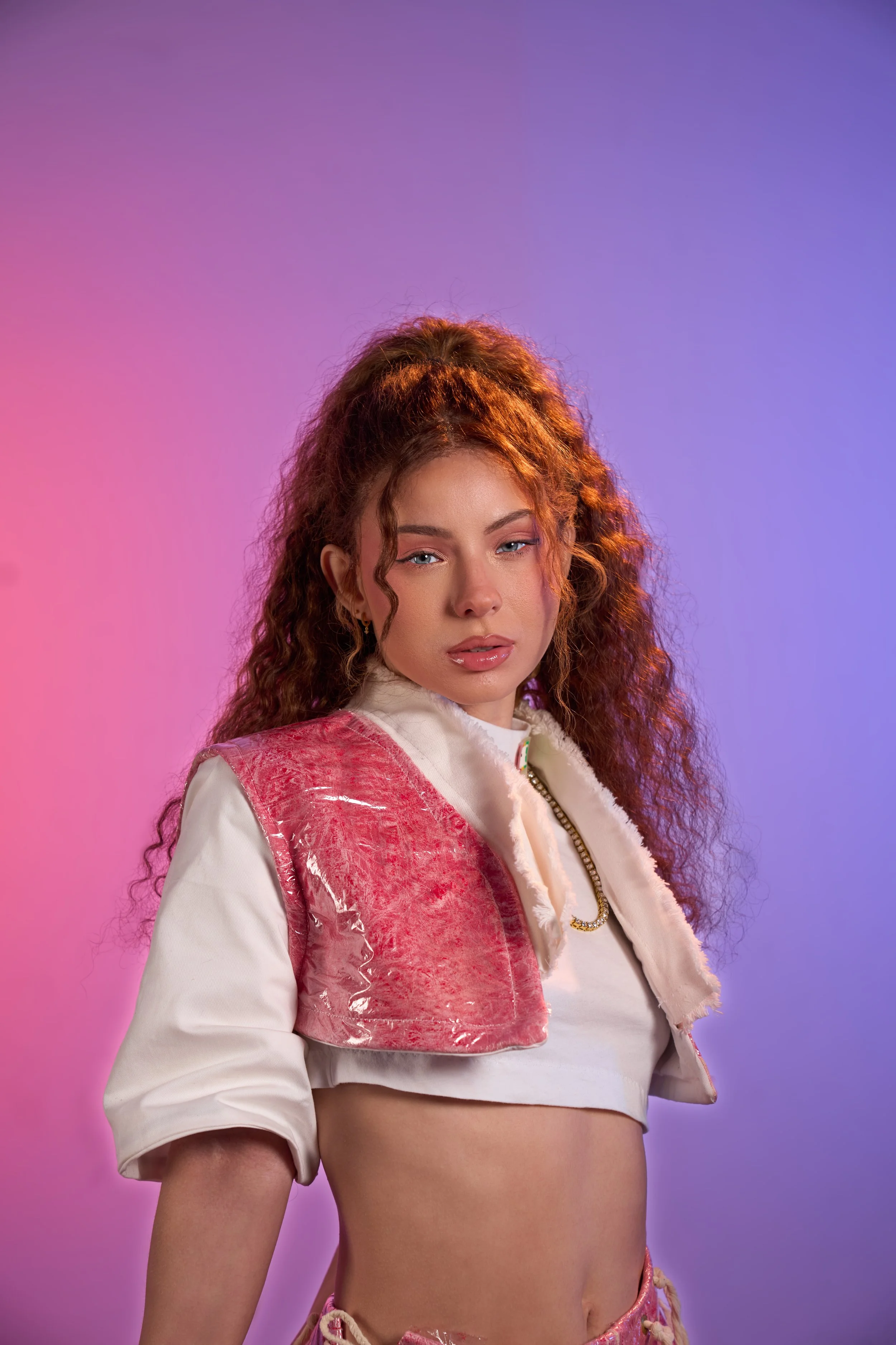 A young woman with curly red hair wearing a cropped white top and a pink vest, standing against a pink and purple gradient background.