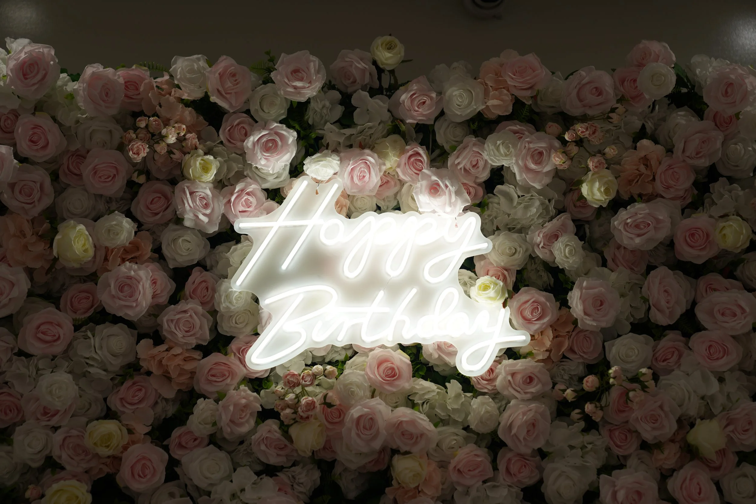 A floral backdrop of pink, white, cream, and peach roses with a neon sign that reads "Happy Birthday" in cursive, surrounded by flowers.