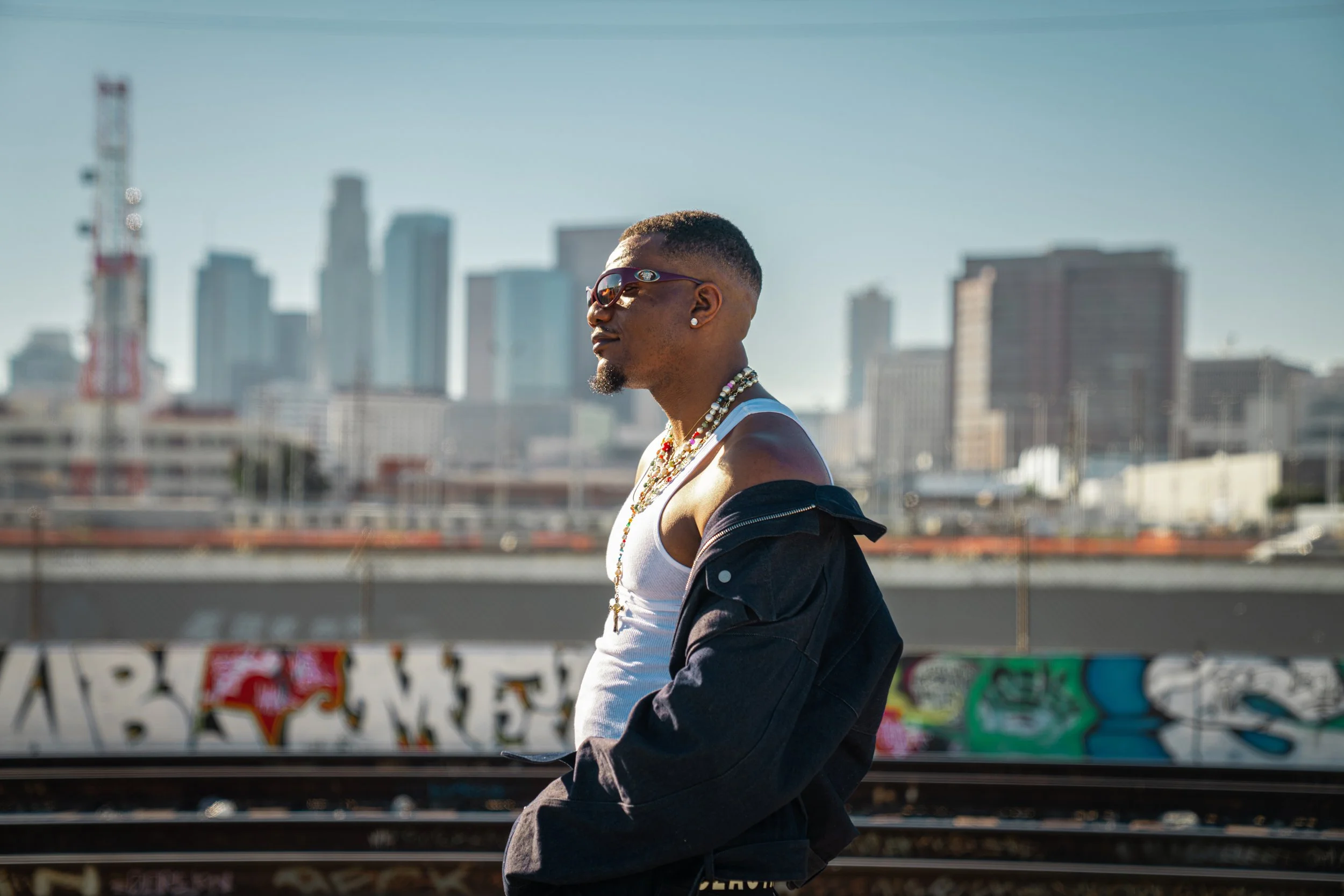 A man with sunglasses and jewelry standing outdoors with a city skyline in the background.
