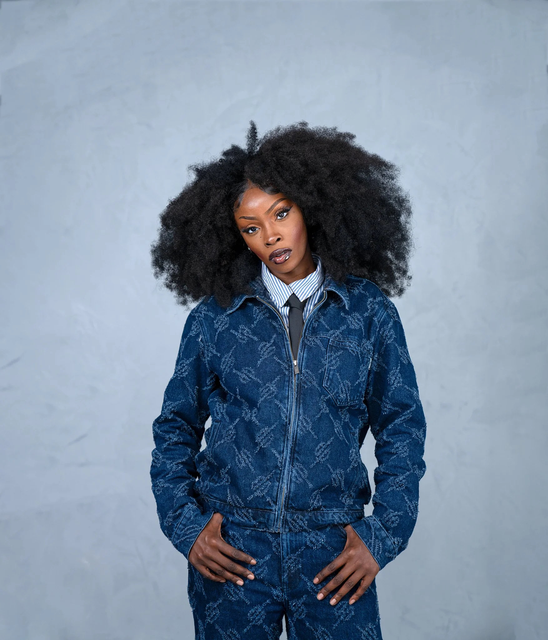 A woman with dark skin and large curly black hair wearing a blue patterned jacket over a striped shirt and tie, standing against a plain gray background.