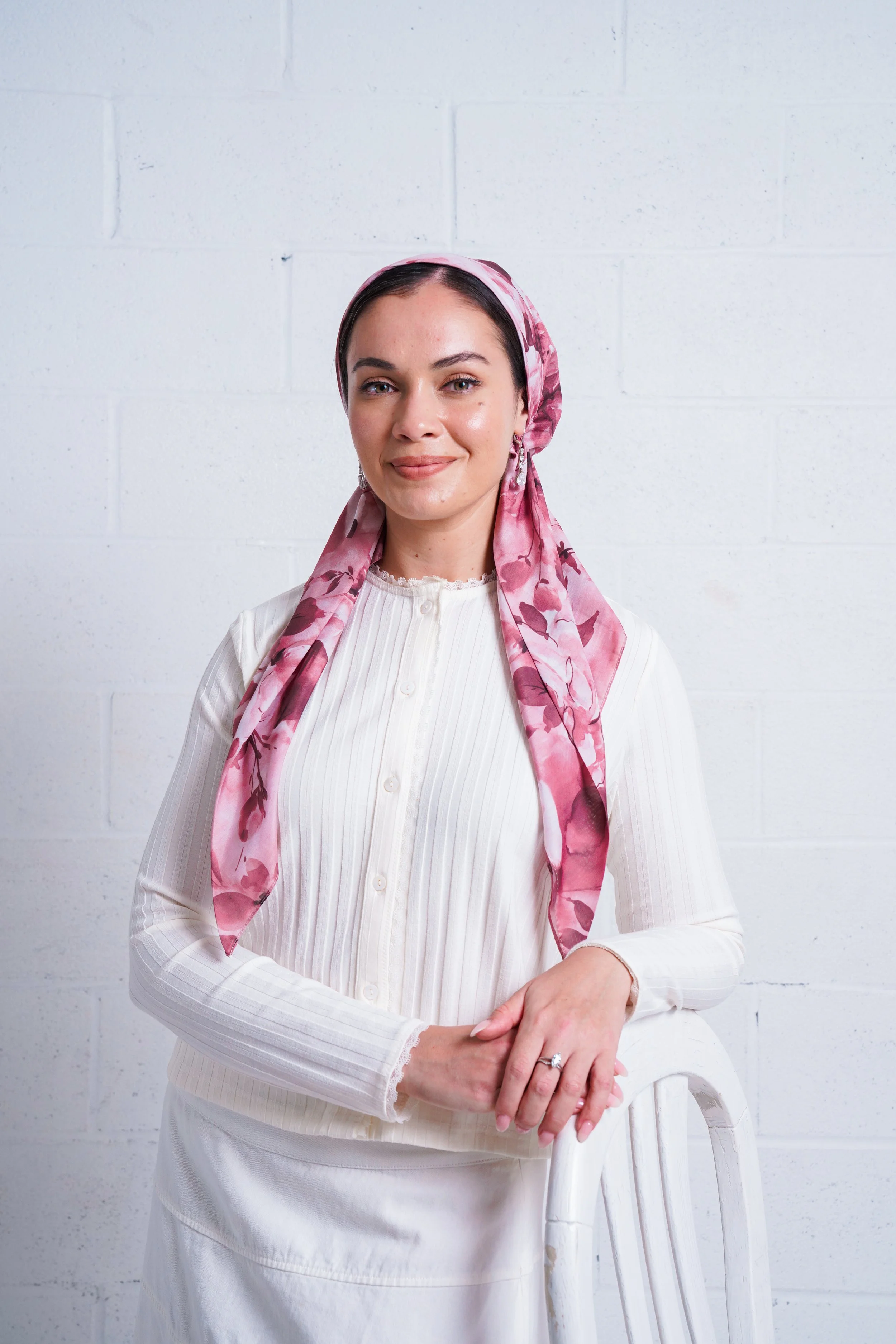 A woman with light skin, dark hair, and brown eyes, wearing a pink floral headscarf and a white long-sleeve blouse, standing against a white brick wall.