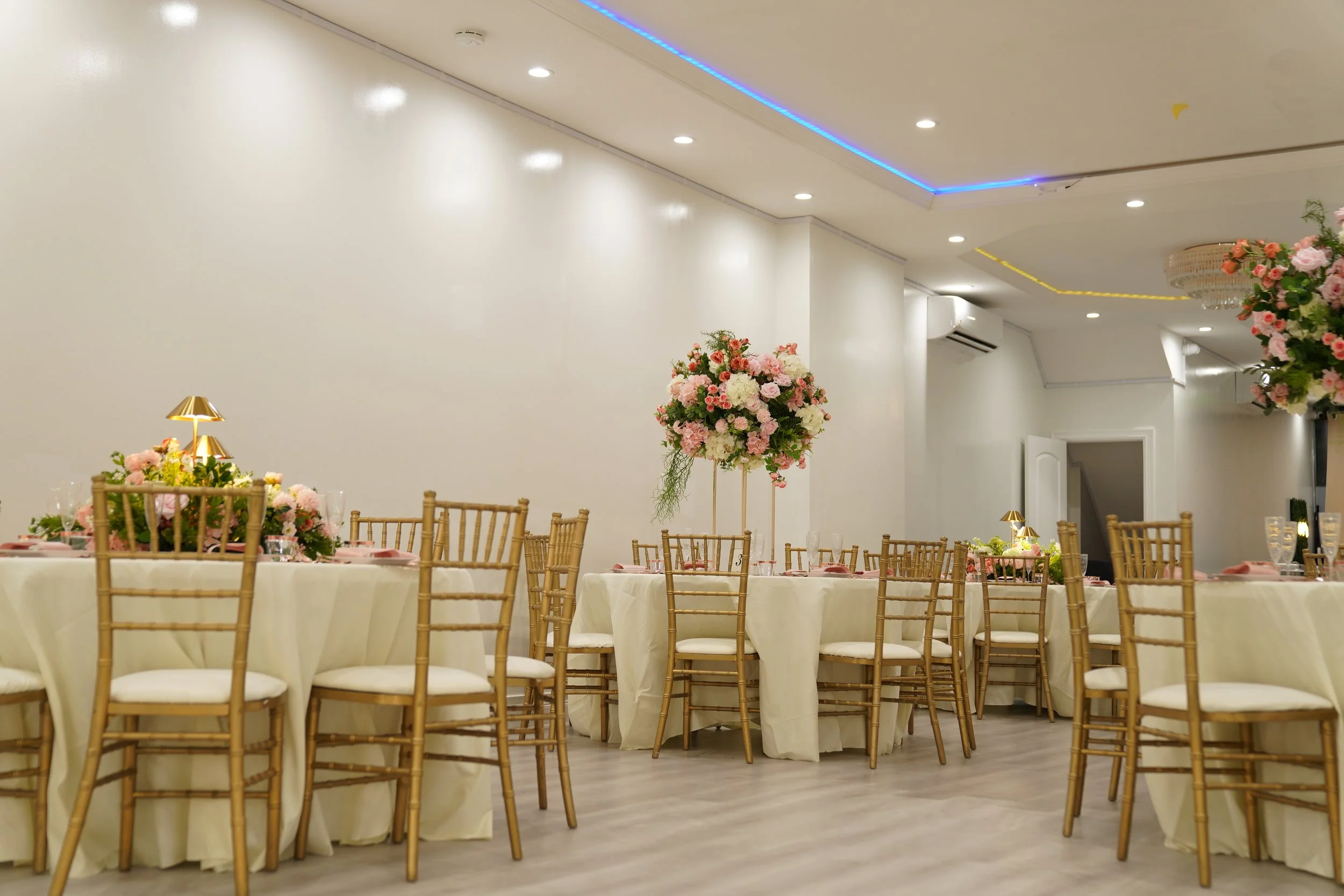 Elegant banquet hall decorated with pink and white floral arrangements on round tables draped in white tablecloths, surrounded by wooden chairs with white cushions, and accented with gold tableware and lamps.