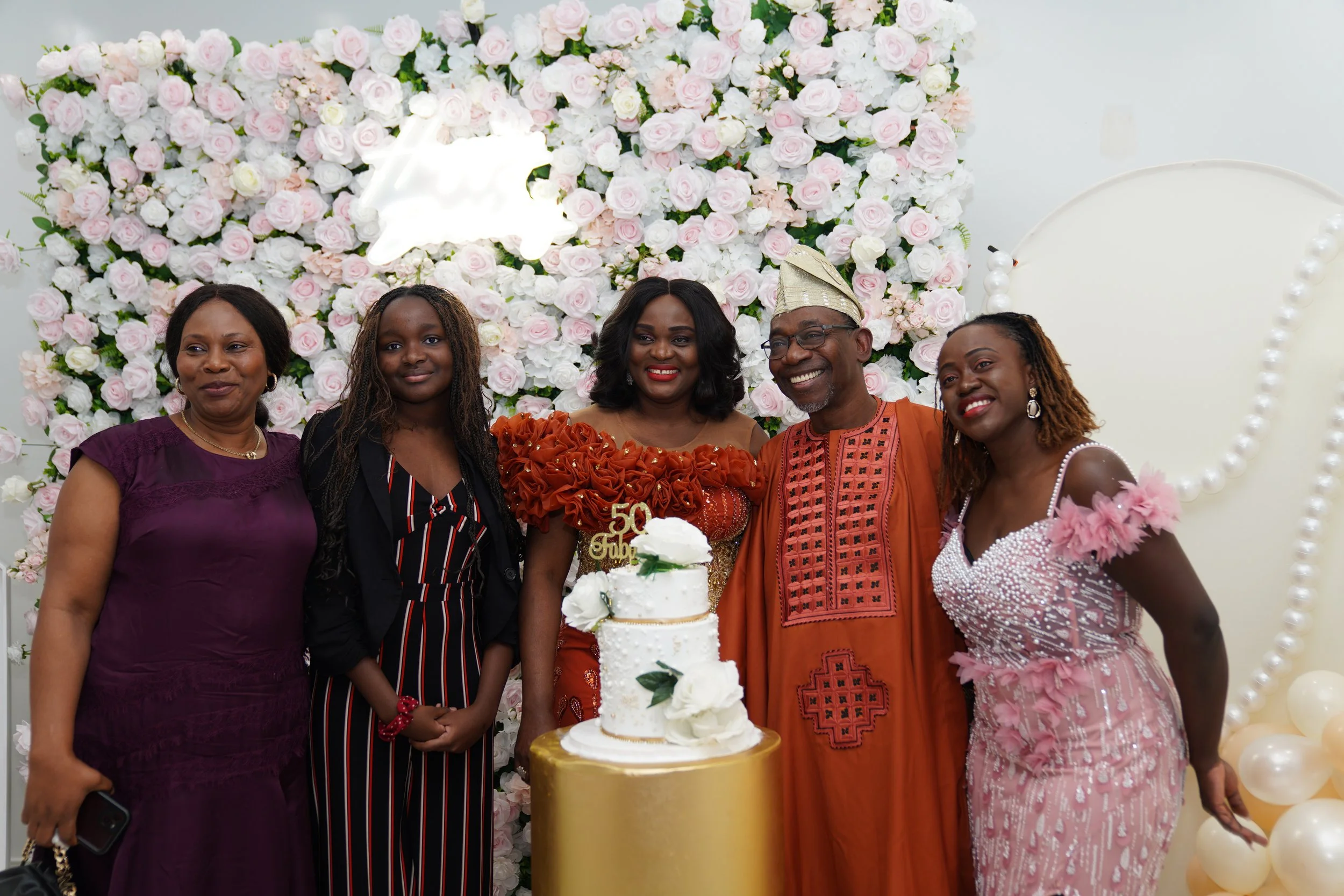 A group of six people celebrating a 50th birthday in front of a floral backdrop with pink and white roses, with a cake decorated with white flowers and a gold "50th birthday" topper.